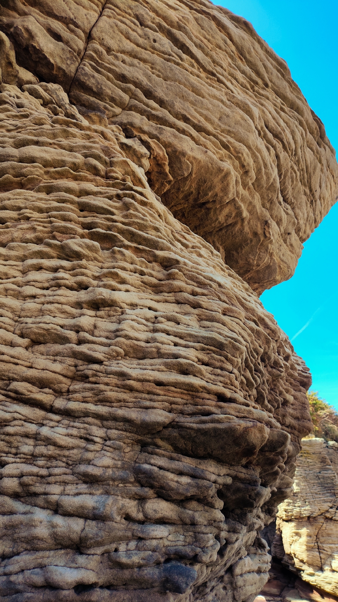 The image shows a close-up, low-angle view of weathered sandstone rock formations against a bright blue sky. The rocks exhibit distinct horizontal layering and erosion patterns, creating a textured and rugged surface. The formations are predominantly brown and tan in color, with some darker, shadowed areas. In the background, on the right side of the frame, another rock formation with similar characteristics is visible, along with a hint of green foliage at the very top right. The scene is devoid of any people or specific urban structures, suggesting a natural or coastal environment. This photograph is taken at Red Rocks Nature Reserve in West Kirby, United Kingdom, a location known for its extensive red sandstone cliffs and coastal scenery.