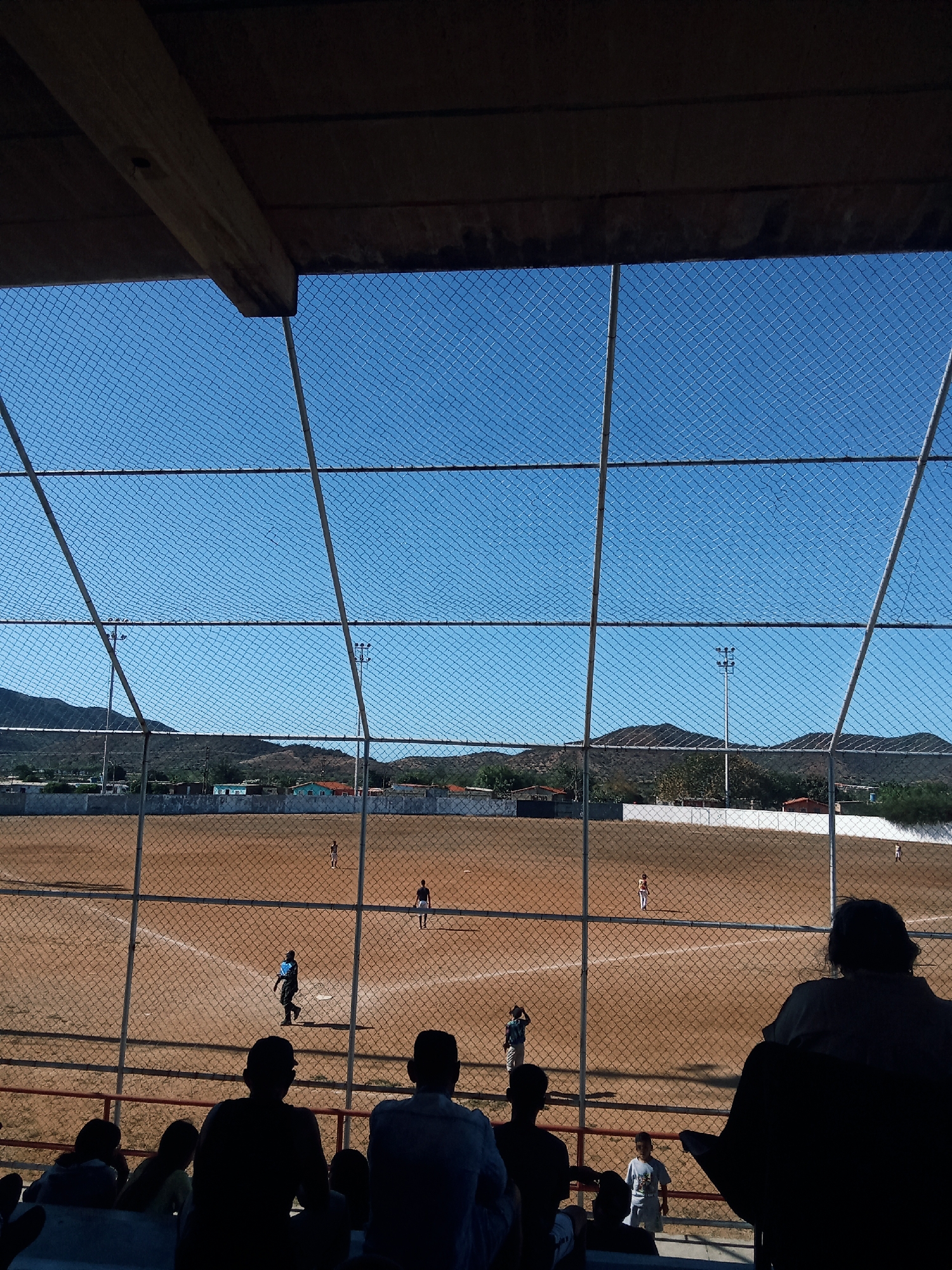 From what appears to be a covered seating area, a baseball field can be seen through a chain-link fence. There are several people on the field. The audience in the seating area is silhouetted. In the background are hills and structures that suggest that the location is in the city of Juan Griego, Venezuela. The sky is clear and blue, and it appears to be daytime.