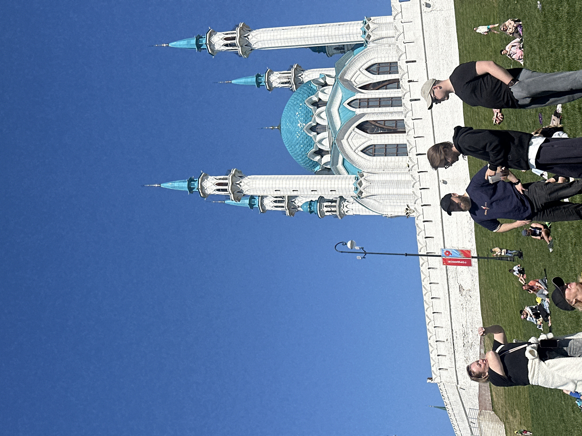 The image features the Kul Sharif Mosque in Kazan, Russia. The mosque is visible against a clear blue sky. Adjacent to the mosque is a grassy area with several people. Some are standing and walking, while others are lying down. A white wall extends along the edge of the grass, and a street lamp stands nearby.