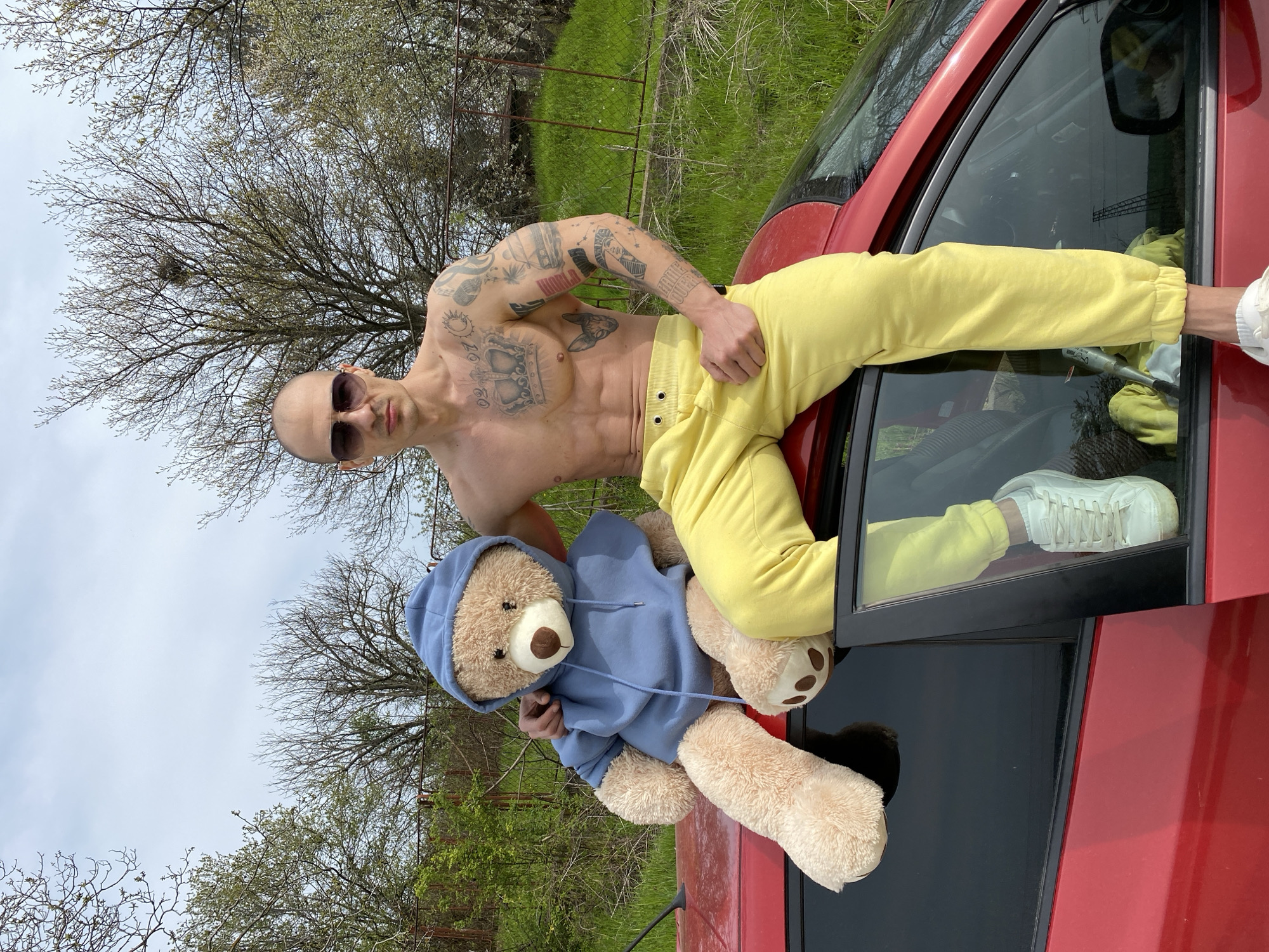 A shirtless man with sunglasses and yellow pants poses on a red car next to a teddy bear wearing a blue hoodie. The man is muscular and has multiple tattoos on his arms and chest. The car is parked in a grassy area with trees in the background. The location appears to be outdoors, potentially on the outskirts of Pleven, Bulgaria, given the environment depicted. The man is looking directly at the camera, while the teddy bear is positioned next to him. The man's leg is inside the car window and his arm is resting on the car's roof. The teddy bear's arm is also in the window.