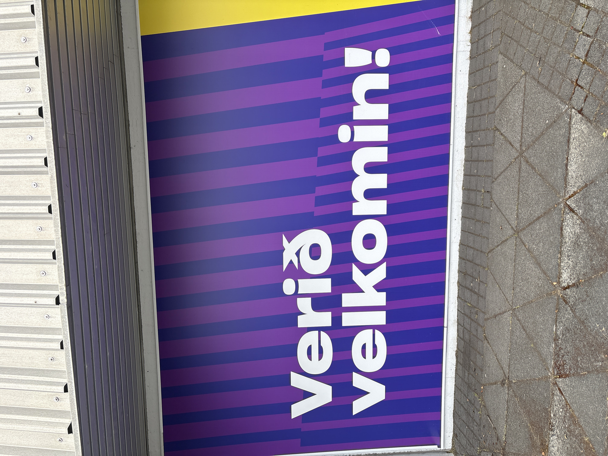 A white text sign reads "Verið velkomin!" against a purple and blue striped background with a yellow vertical stripe on the right. The sign is mounted on the exterior wall of a building, above a paved walkway. The building's facade features corrugated metal siding at the top and a dark grey overhang. The walkway is made of square concrete pavers arranged in a diamond pattern.