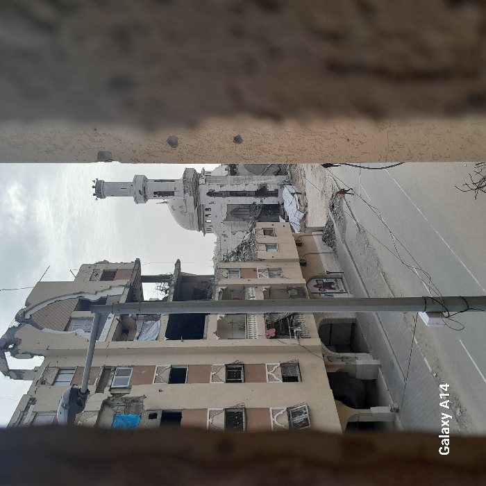 An image shows a war-torn street with damaged buildings and a mosque in the background. The buildings are severely damaged, with exposed interiors and rubble. A tall, white mosque with a dome and minaret is visible amidst the devastation. The street is visible, and there are signs of destruction everywhere. The picture is taken from an elevated position, with a balcony or window ledge framing the bottom. The photograph appears to be taken with a Galaxy A14.