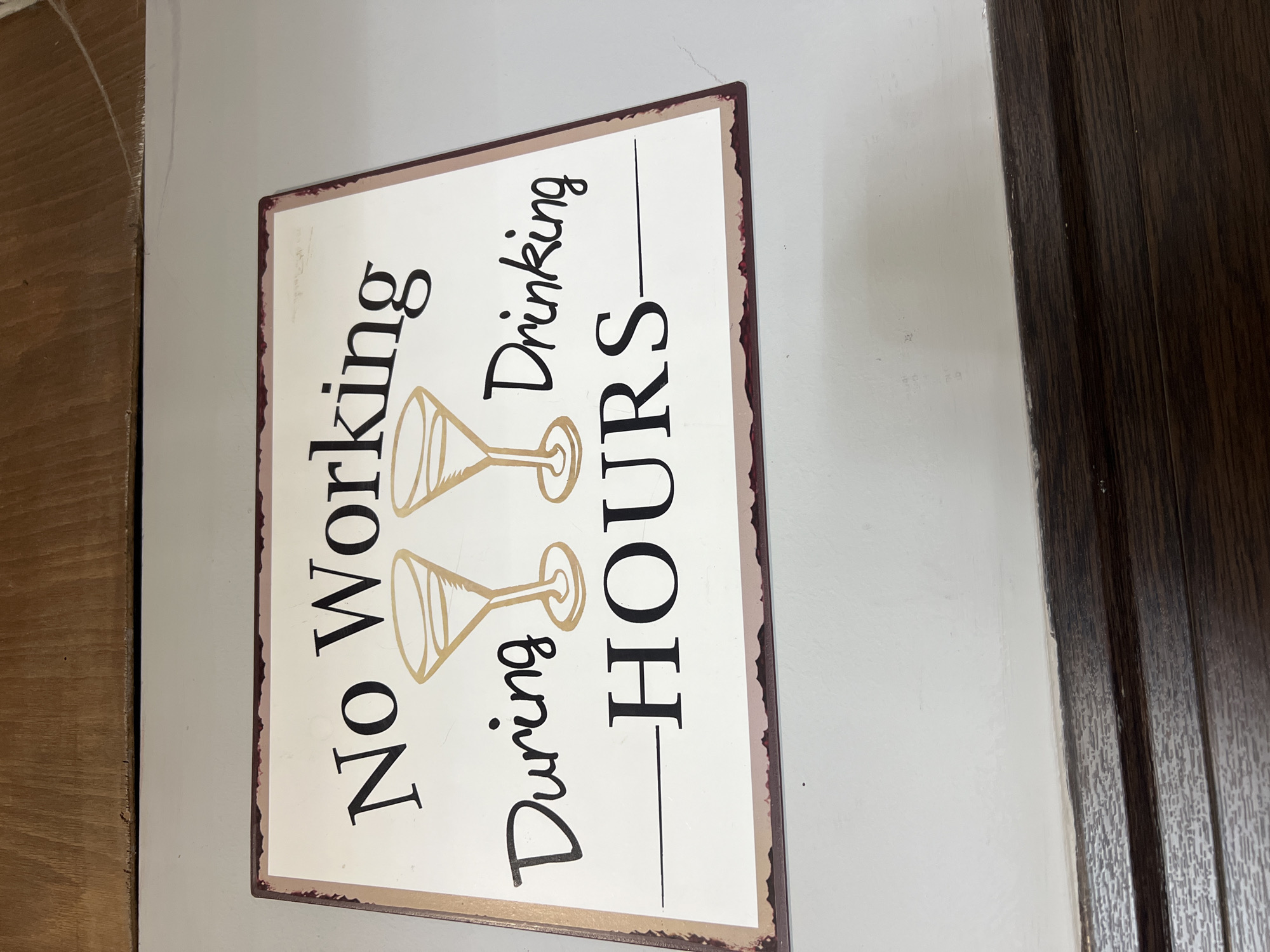 Here is a forensic description of the image:
The image shows a rectangular metal sign with a white background and a faux-distressed dark brown border. The sign contains the text "No Working" stacked vertically, followed by two images of martini glasses. Below the martini glasses is the text "During Drinking," stacked vertically, and finally, the text "HOURS," with a horizontal line above and below. The text is in black, and the martini glasses are in gold. The sign is hanging on a white wall, next to a dark brown wooden door frame. The location is unknown.