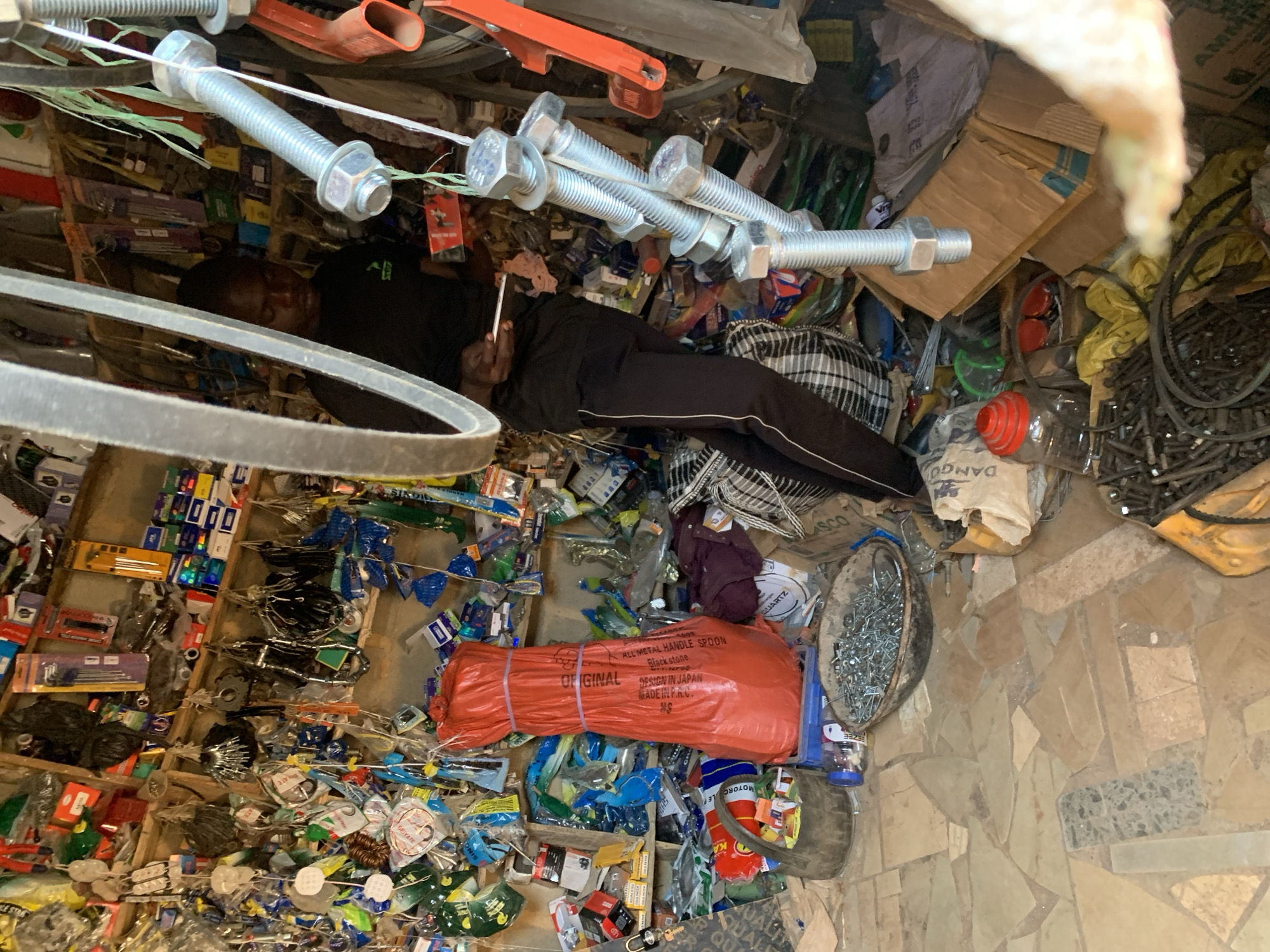 An overhead shot captures the cluttered interior of a small, informal shop or storage area in Gura Top, Nigeria, filled with hardware supplies and various goods. An adult male is lying on his side in the center of the frame, resting amidst the merchandise. He is dressed in dark clothing, including black pants with a white stripe down the side, and is holding a thin, light-colored object, possibly a cigarette.
The scene is densely packed with items, including shelves and various surfaces laden with small packages, tools, and hardware. Large bolts and nuts are visibly hanging on a string near the top of the frame. Other items include coils of wire or belts, bowls filled with nails or screws, cardboard boxes, and plastic-wrapped products. A large, orange-red package, possibly a tarp or bulky item, is prominent in the lower middle. The floor is covered with irregularly shaped, light-colored tiles.
Visible text in the image includes: "SEIYATE", "ALL METAL HANDLE SPOON", "Black stone", "ORIGINAL DESIGN IN JAPAN", "MADE IN P.A.C.", "HS", "MOTORC", "REZEE", "QUARTZ", "ASCO", "ODNVA", and "AMMA". The chaotic arrangement suggests a busy commercial environment with a wide variety of stock.