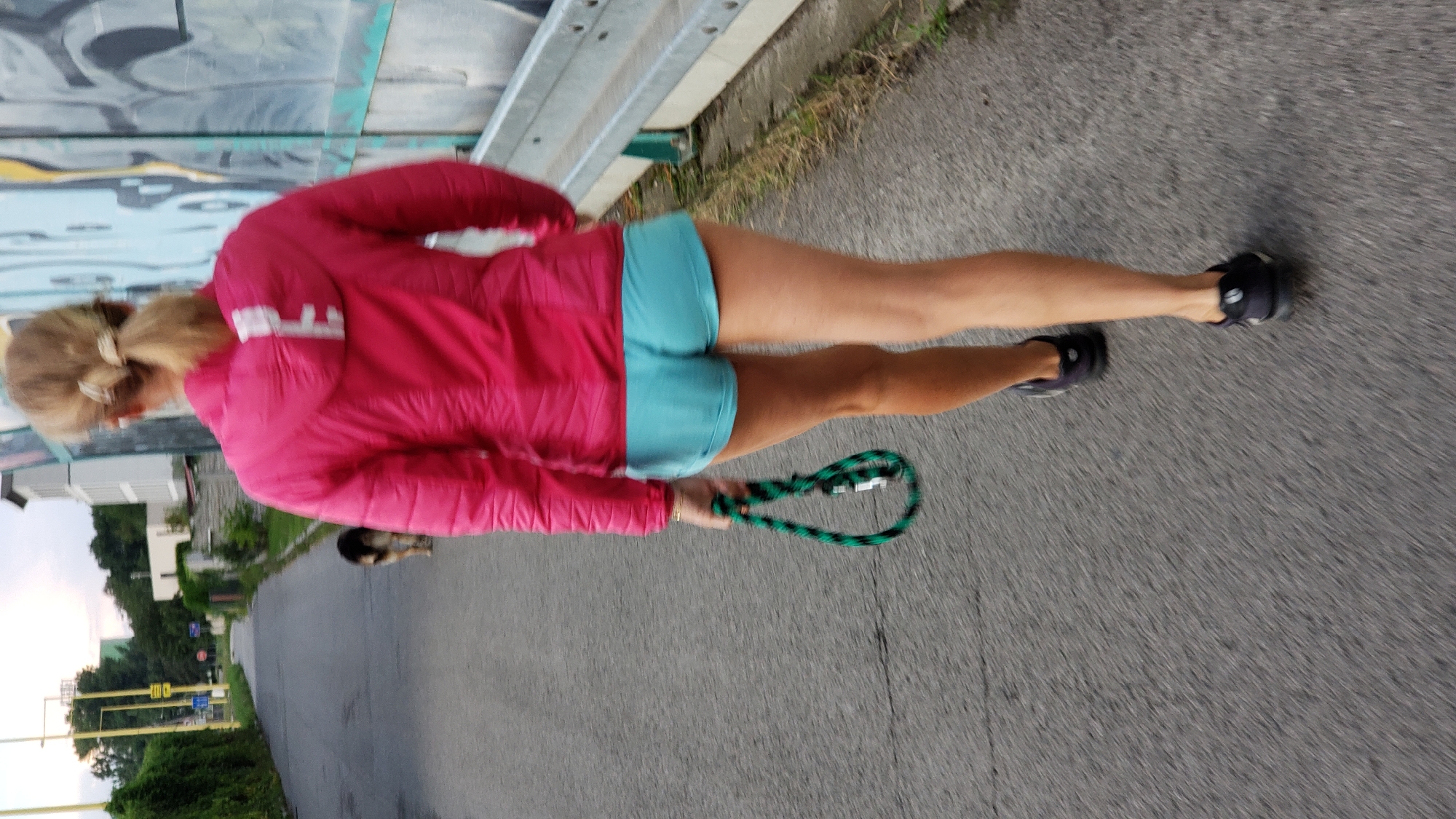 A woman with blonde hair in a ponytail, wearing a pink puffer jacket and light blue shorts, walks on an asphalt road. She is holding a green and black braided leash. A dog, possibly a German Shepherd, walks ahead of her. The scene takes place in an urban environment with buildings and graffiti visible in the background. The location is identified as Prešov, Slovakia. The lighting suggests it is late afternoon or early evening.