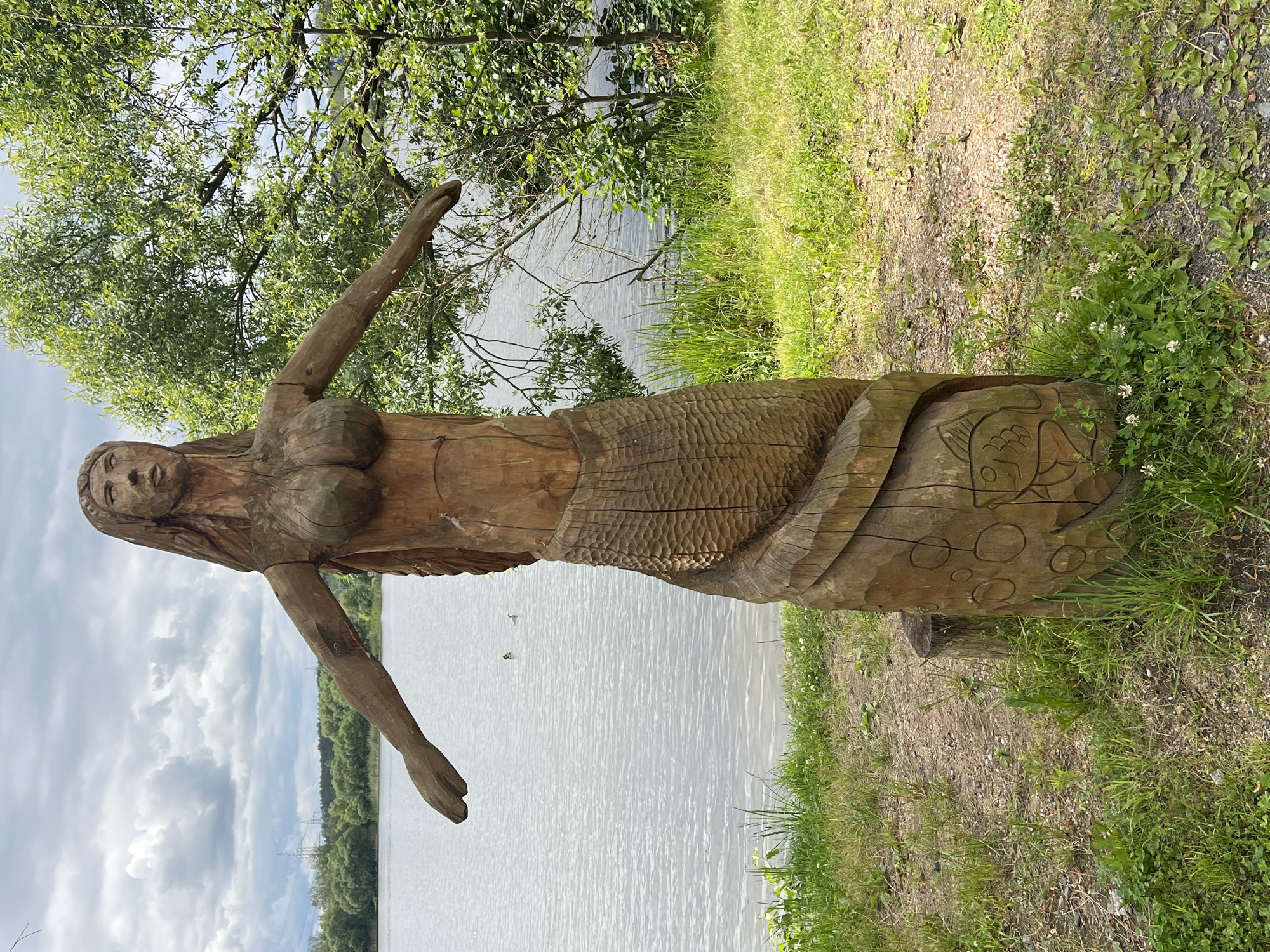 A wooden sculpture of a mermaid stands on the grassy bank of a lake. The mermaid is carved from a single log and has her arms outstretched. Her lower body is carved to resemble a fish tail, and there are carved circular patterns and a fish design on the tail. The background consists of a calm lake with a few birds swimming on it, and lush green trees and vegetation along the opposite bank. The sky is partly cloudy with some visible power lines in the distance. The sculpture is located near the Minsk Sea (Zaslavskoye Reservoir), a popular recreational area outside of Minsk, Belarus.