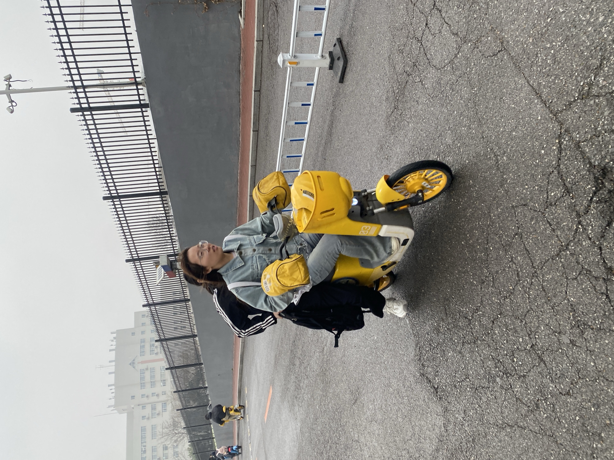 A young woman in a denim jacket and jeans is riding a yellow electric scooter on an asphalt surface. She has a yellow bag strapped across her body and a black backpack at her feet. She wears white sneakers and glasses. A black metal fence runs parallel to the road, with a gray wall behind it. In the background, tall buildings are visible through the hazy weather, and a ladder is visible on the right side of the road. Other people riding similar scooters are visible in the distance. The photo was likely taken in the city of Zhongtun, China.