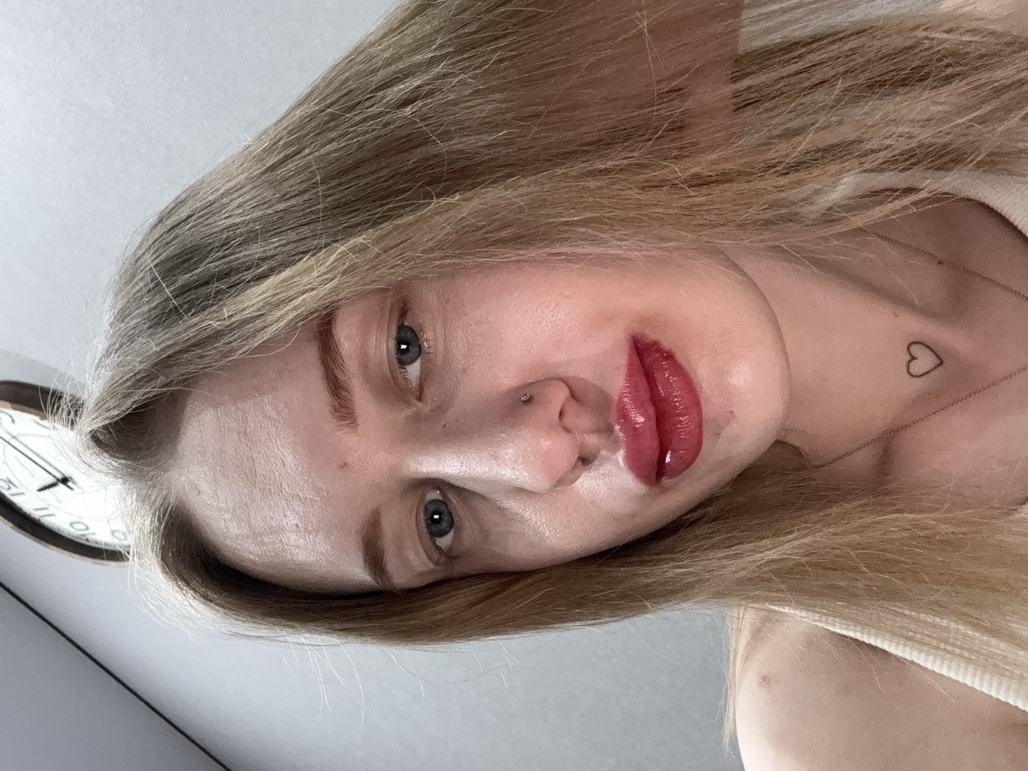 A close-up, eye-level indoor photograph depicts a female subject, frontal view, occupying the majority of the frame. The subject has fair skin, blonde hair parted on her left, blue eyes, and full lips coated in a glossy, dark red substance. A small, silver-colored stud is present in her left nostril. A faint, black outline of a heart is tattooed on the left side of her neck, above a thin, gold-colored chain. She wears a white ribbed tank top. A circular wall clock with a white face and black numerals indicating approximately 1:30 is partially visible in the upper right background. The background consists of a plain, light grey wall. The scene is situated in an interior location in Rostov-on-Don, Russia.