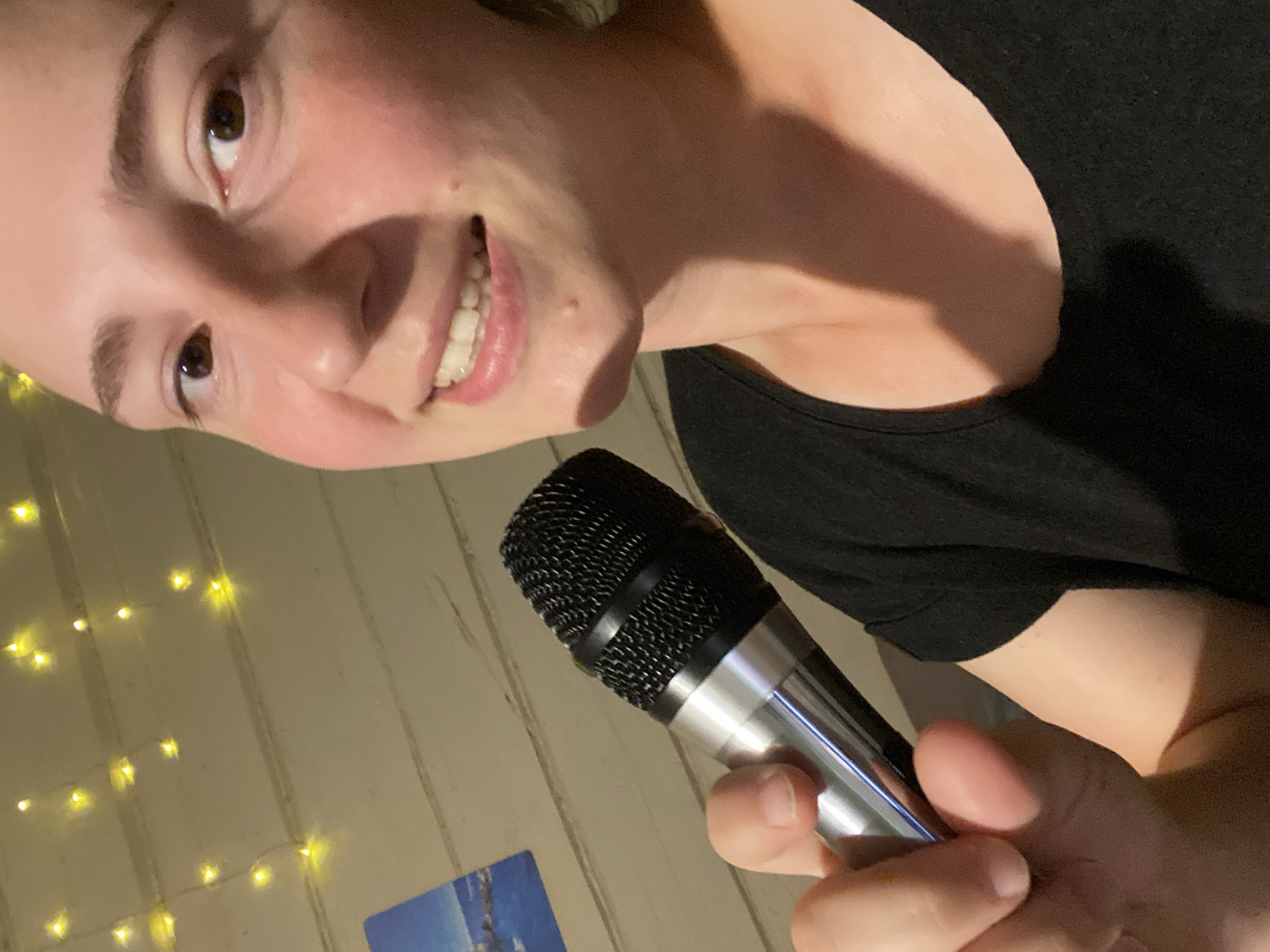 A young woman is holding a microphone in her right hand, close to her face. She is wearing a black t-shirt. Her head and shoulders are visible, and she is smiling with her teeth showing. The background is a wooden wall with string lights. The lighting is dim, with a warm glow from the string lights.