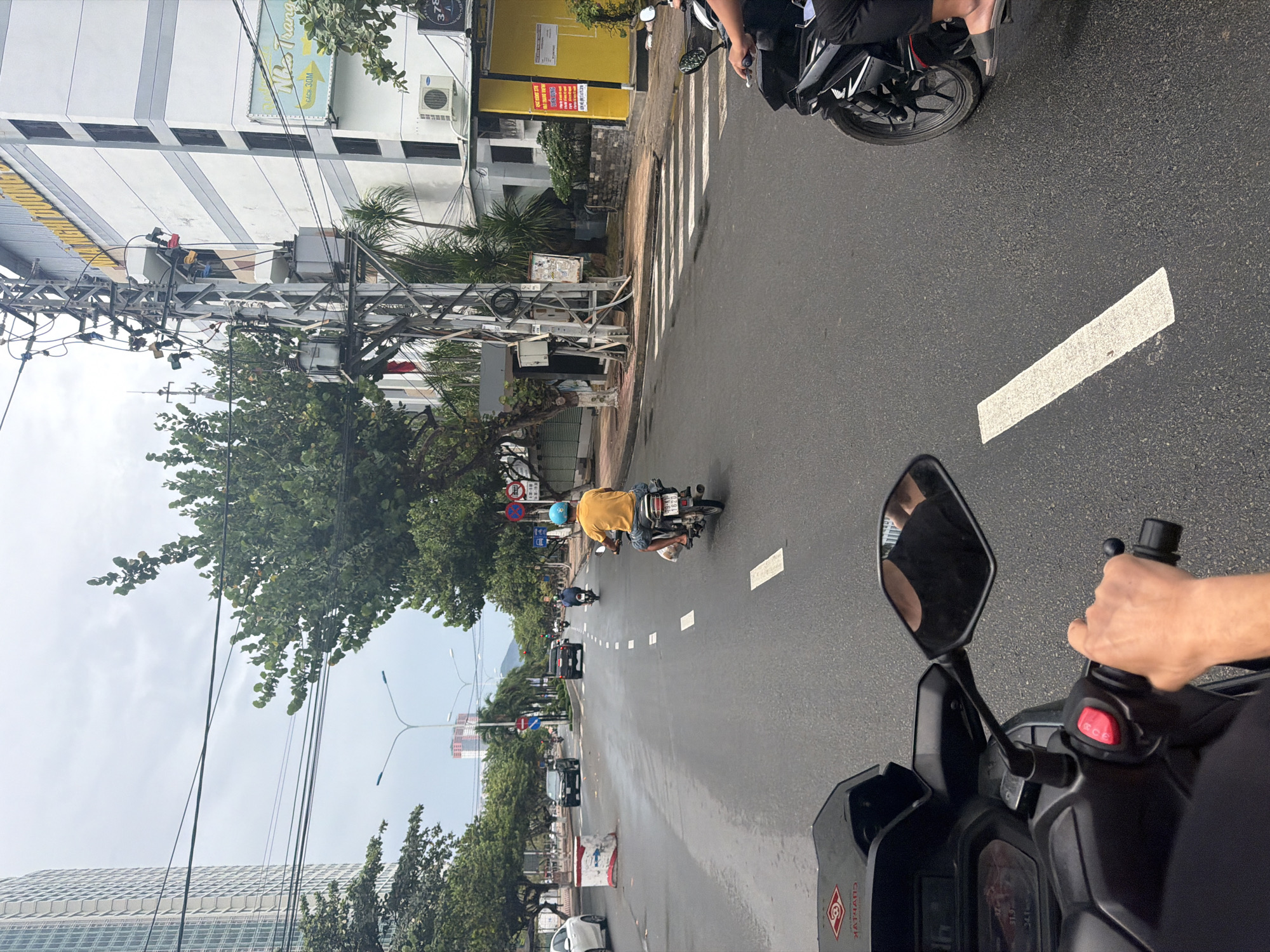 The image depicts a street scene in Nha Trang Ward, Vietnam, taken from the perspective of a person riding a motorcycle. In the foreground, the motorcycle's handlebars, mirror, and rider's hand are visible. The asphalt road is wet and marked with white rectangular lane dividers. Several other motorcycles are present on the road, some with riders visible. Buildings line the left side of the street, including a white building with air conditioning units and power lines running along the side. A crosswalk is marked on the road near the buildings. Trees are present on both sides of the road, partially obscuring views of distant buildings, including a high-rise structure. The sky is overcast.