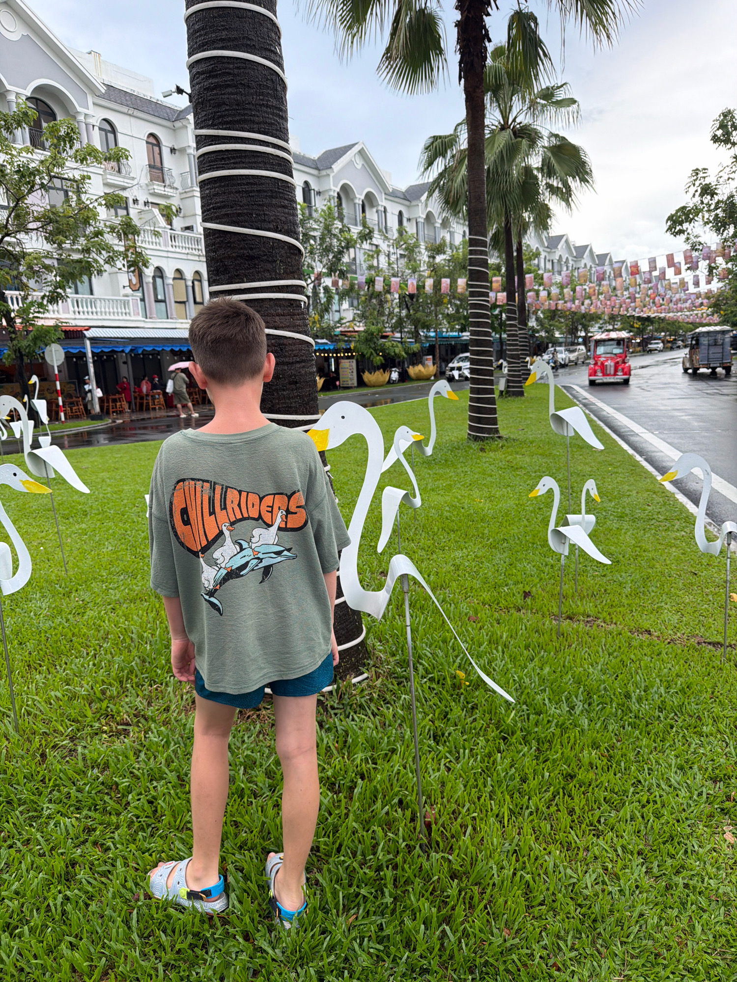 In the city of Phú Quốc, Vietnam, a young boy stands in a grassy area, facing away from the camera. He is wearing a green t-shirt with a graphic design on the back featuring text that appears to read "CHILL RIDERS" above stylized images of ducks and a dolphin. He is also wearing blue patterned shorts and sandals. The boy is positioned in front of a large palm tree that has white stripes wrapped around its trunk. Scattered around the boy and the palm tree are several white metal sculptures resembling swans with yellow beaks. In the background, a street is visible with cars and a red open-air vehicle. Buildings with white facades and gray roofs line the street, and colorful banners are strung across it. The overall atmosphere suggests an outdoor public space with decorative elements.