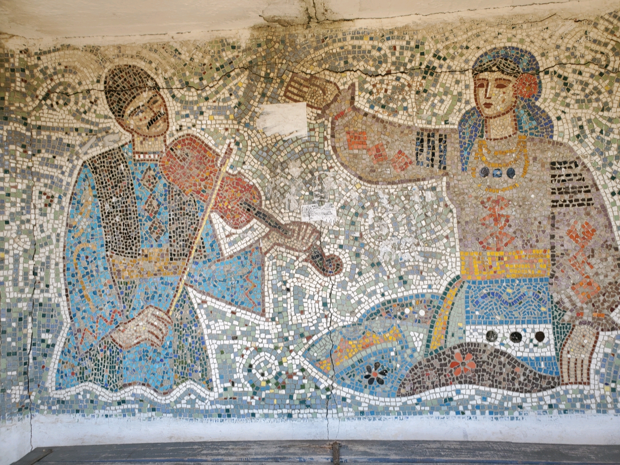 This is a mosaic mural depicting two figures. On the left, a man with a dark hat and mustache plays a violin. He is dressed in a blue garment with intricate patterns. On the right, a woman with blue hair, adorned with a flower, wears a light-colored dress with yellow and blue accents. She has a necklace with blue pendants. The figures are set against a background of abstract, wave-like patterns in various shades of green, grey, and white. The mosaic shows signs of age and damage, with visible cracks running through it. The mural appears to be located outdoors, possibly on a wall or building facade, with a light-colored ceiling visible at the top. Below the mural is a dark, horizontal surface. This mosaic is located in Pervomaisk, Moldova.