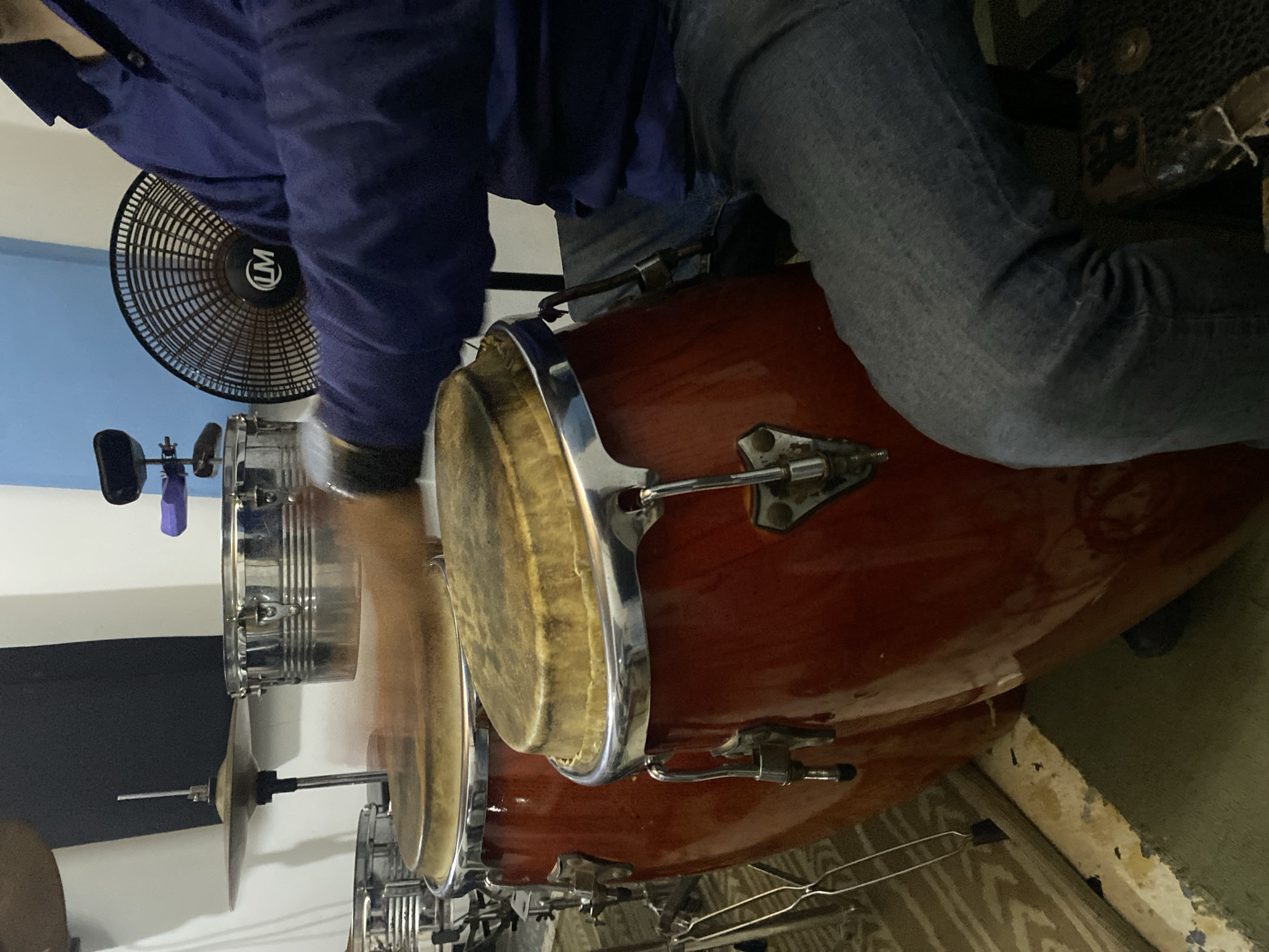 A person wearing jeans and a blue shirt is seated, with their leg resting against a red conga drum. The drum set is in what appears to be a room with damaged walls and what may be a concrete or painted floor. An electric fan is visible in the background, positioned against a wall that is painted in different shades of blue and white. Part of a drum set, including cymbals and other percussion instruments, is also in the frame. The location is Tinaquillo, Venezuela.