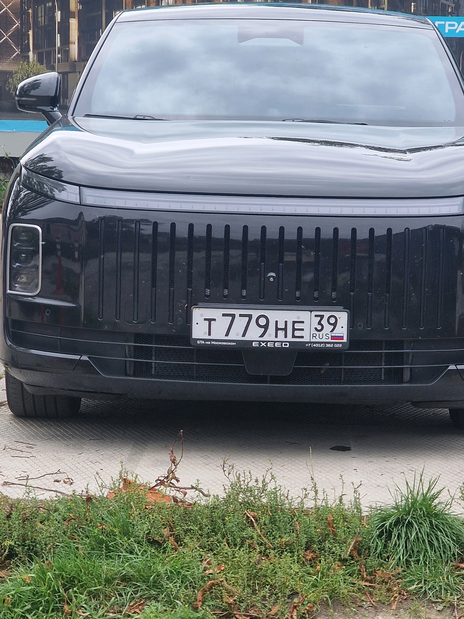 The image shows the front of a black EXXEED car parked on a paved surface with grass in the foreground. The car has a license plate that reads "T 779 HE 39". Below the license plate, there is text that reads "EXXEED" and "+7 (4012) 352 022". The background shows a building with a blue sign on it. The scene is in Zelenogradsk, Russia.