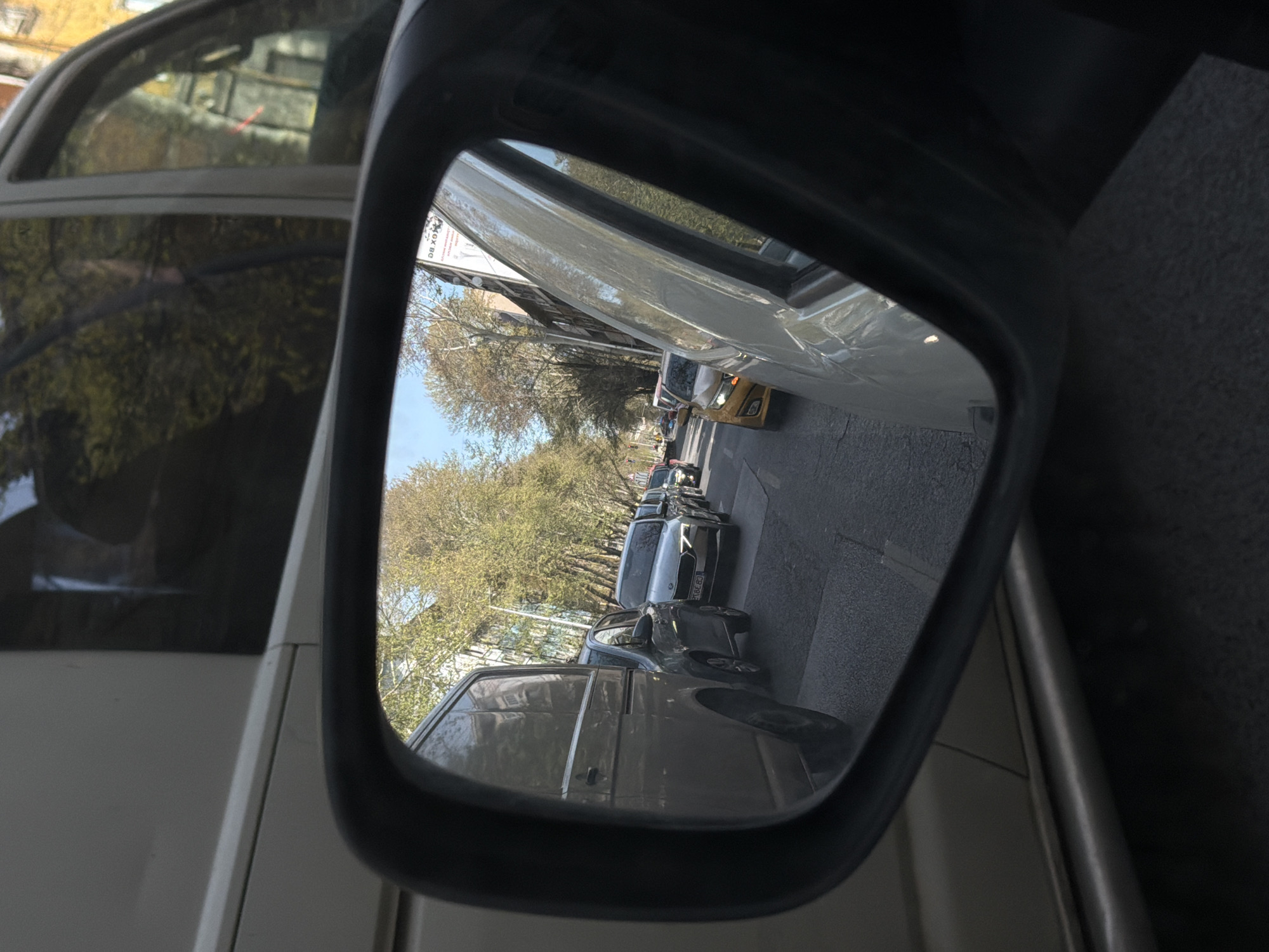 The image displays a vehicle's side mirror, which is centrally framed and appears vertically oriented within the rotated photograph. The reflection inside the mirror shows a street scene laid out horizontally.
Within the reflection, a line of parked cars extends into the distance along what appears to be a curb or sidewalk. Above the vehicles, numerous trees with sparse, light green foliage suggest either early spring or late autumn. A clear blue sky is visible above the trees and cars, indicating bright daytime conditions. The scene is well-lit by sunlight.
Various vehicles are visible in the reflection, including several dark-colored sedans, vans, and a distinct yellow car further down the street. On one silver parked car, part of a European-style license plate is clearly visible, reading "90 ED". Another white rectangular sign or billboard in the reflection shows faint dark text, partially discernable as "8Q BC".
The mirror itself has a dark, possibly black, frame. To the left of the mirror, a portion of the white body of the vehicle the mirror is attached to is visible, along with reflections in its window of other trees or structures. No people are present in the image. The setting depicts a typical urban or suburban street.