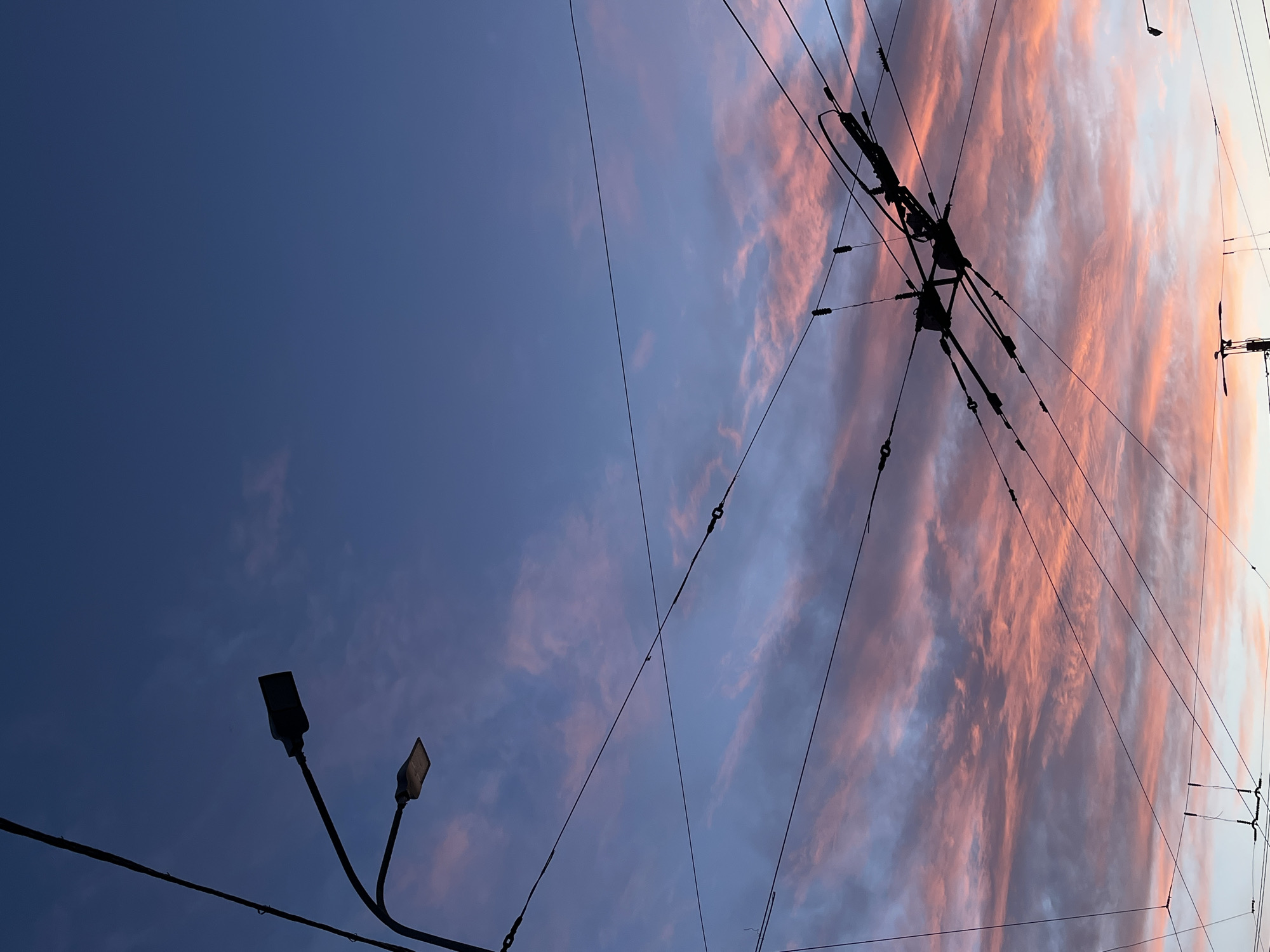 The image captures a view looking upwards in Saint Petersburg, Russia, at dusk or dawn. The sky is a mix of blue and pink-tinged clouds. Silhouetted against the sky are overhead tram or trolleybus power lines and support structures. In the lower left, a street lamp with two light fixtures is visible.