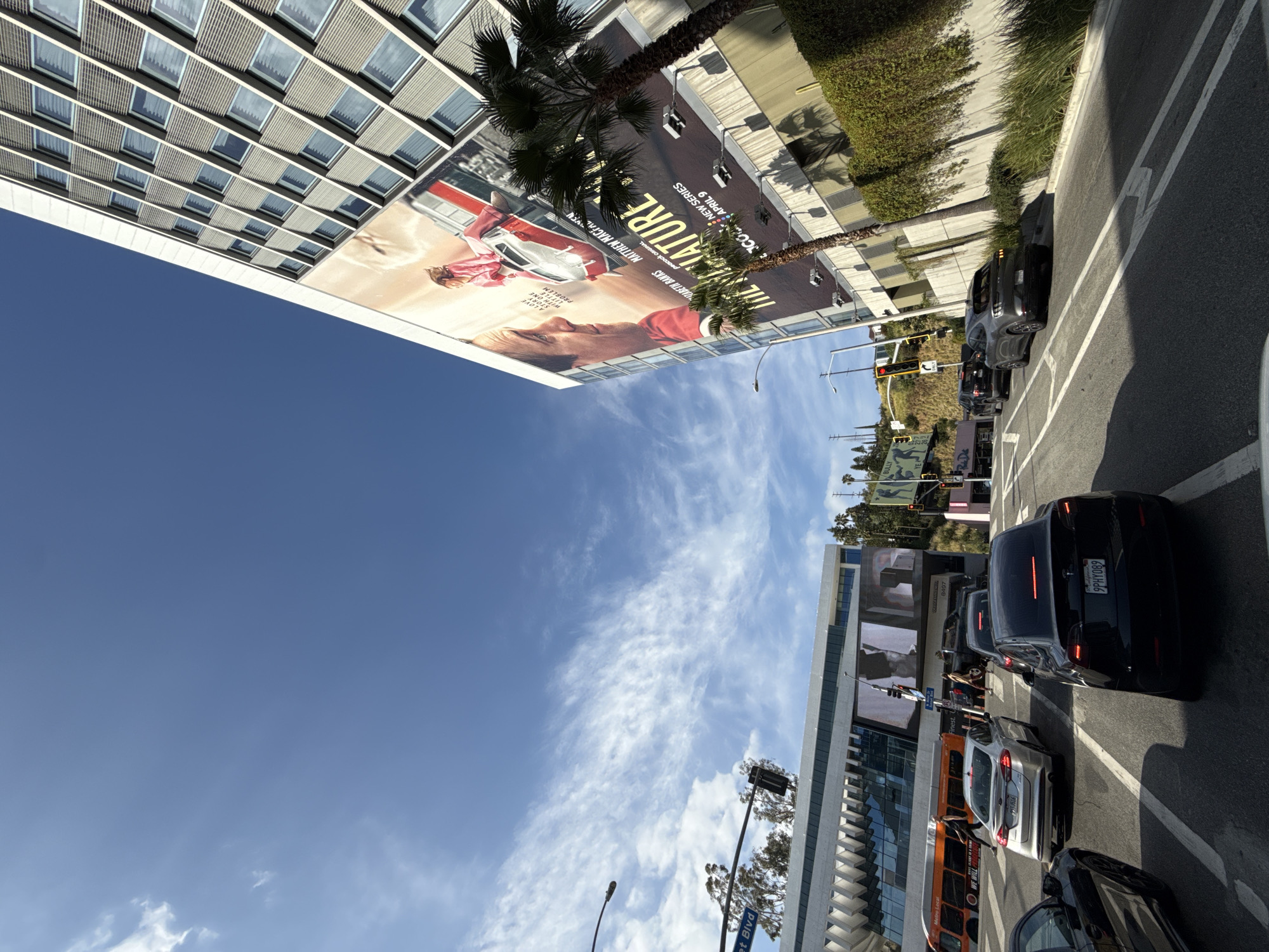 The image shows a street scene in West Hollywood, United States of America. The perspective is from a low angle, looking up at buildings and the sky. On the left is a tall building with many windows and a large advertisement for "The Nature" new series starring Elizabeth Banks and Matthew Macfadyen, premiering April 9. The sky is blue with scattered clouds. On the right is a street with several cars, including a black sedan with license plate "9PHY089" and an orange bus. Street signs including "et Blvd" and "BULLY STOR" are also visible.