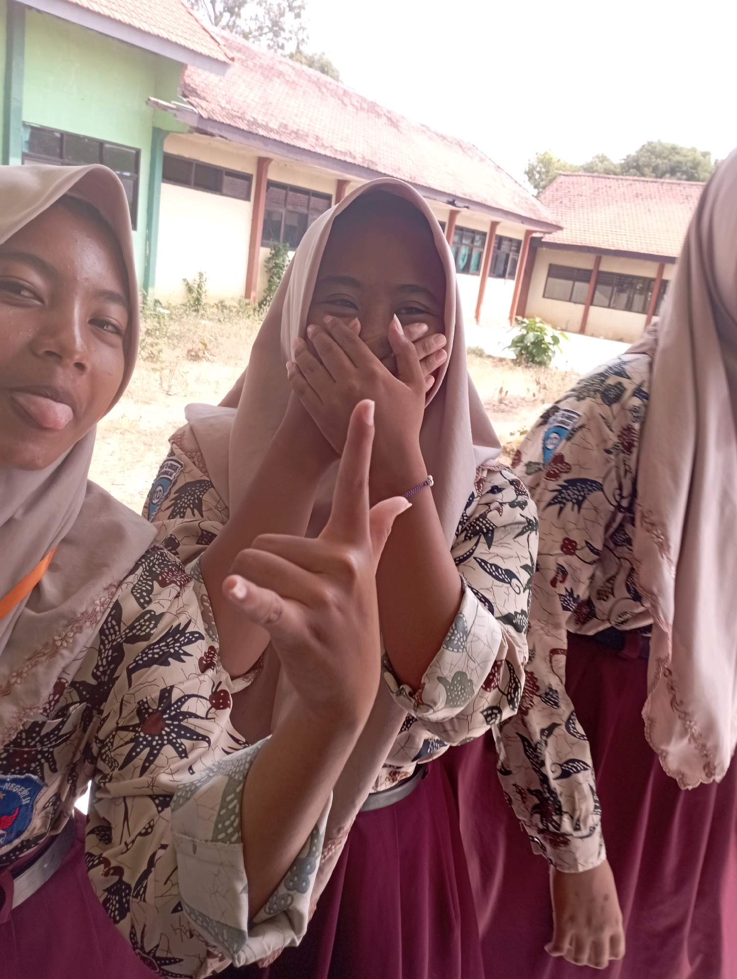Three young people wearing school uniforms are posing for a photo outdoors. The person in the foreground on the left is sticking their tongue out and has their left hand resting on their hijab. The person in the center is covering their mouth with their hands and holding up one finger on their right hand. The person on the right is mostly obscured by the other two. They are in front of a building with a red tiled roof and light-colored walls. The building appears to be a school, possibly located in Banyuurip, Indonesia, given the context. The ground is covered in dry grass and dirt.