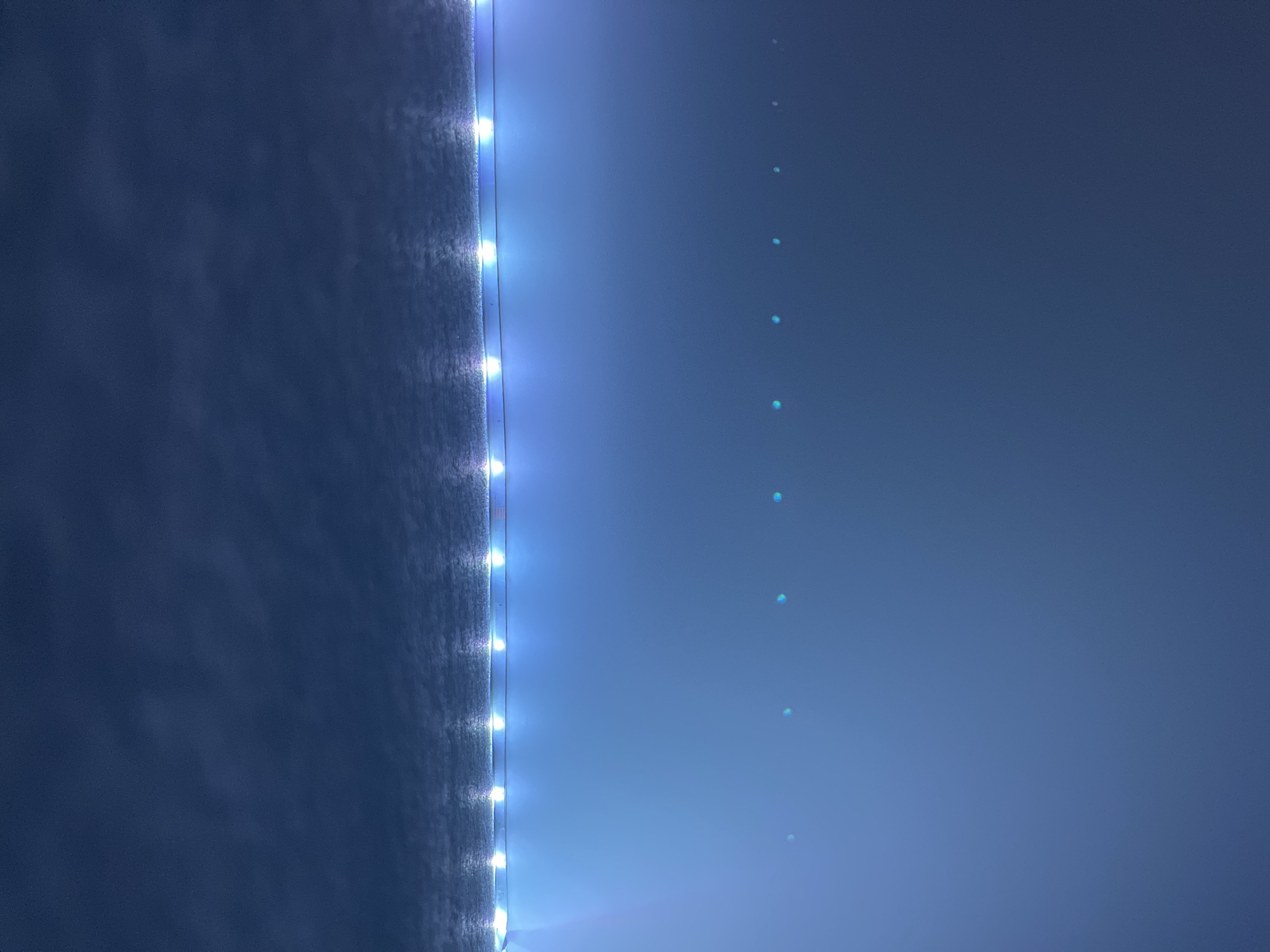 The image shows a wall with a strip of LED lights running vertically along the left side. The wall appears to be textured. To the right of the LED strip is a smooth, blue-toned wall surface. There are small, diffuse spots of light vertically aligned on the right side of the wall. The location is unknown.