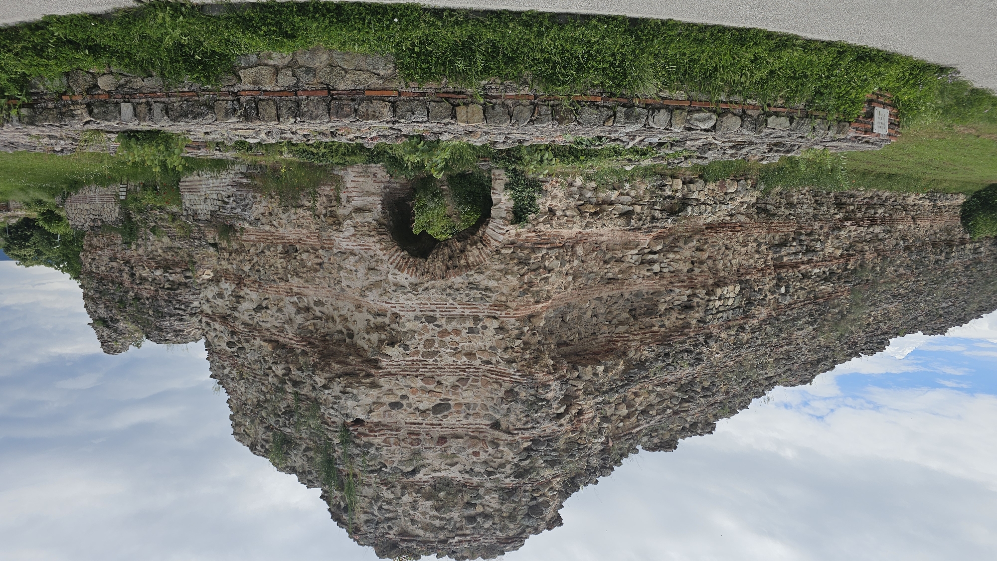 The image depicts an ancient, ruined stone and brick wall, likely part of a fortress, in what appears to be Hisarya, Bulgaria. The wall is constructed of roughly hewn stones interspersed with layers of red bricks. Vegetation, including grass, shrubs, and vines, grows atop and along the wall, indicating weathering and age. Part of the wall has a rounded arched area that is partially filled with vegetation. Above the wall, there is a grassy area with additional stonework bordering it and part of a paved road. The sky is visible in the lower portion of the image, suggesting the wall's height and the surrounding open space.