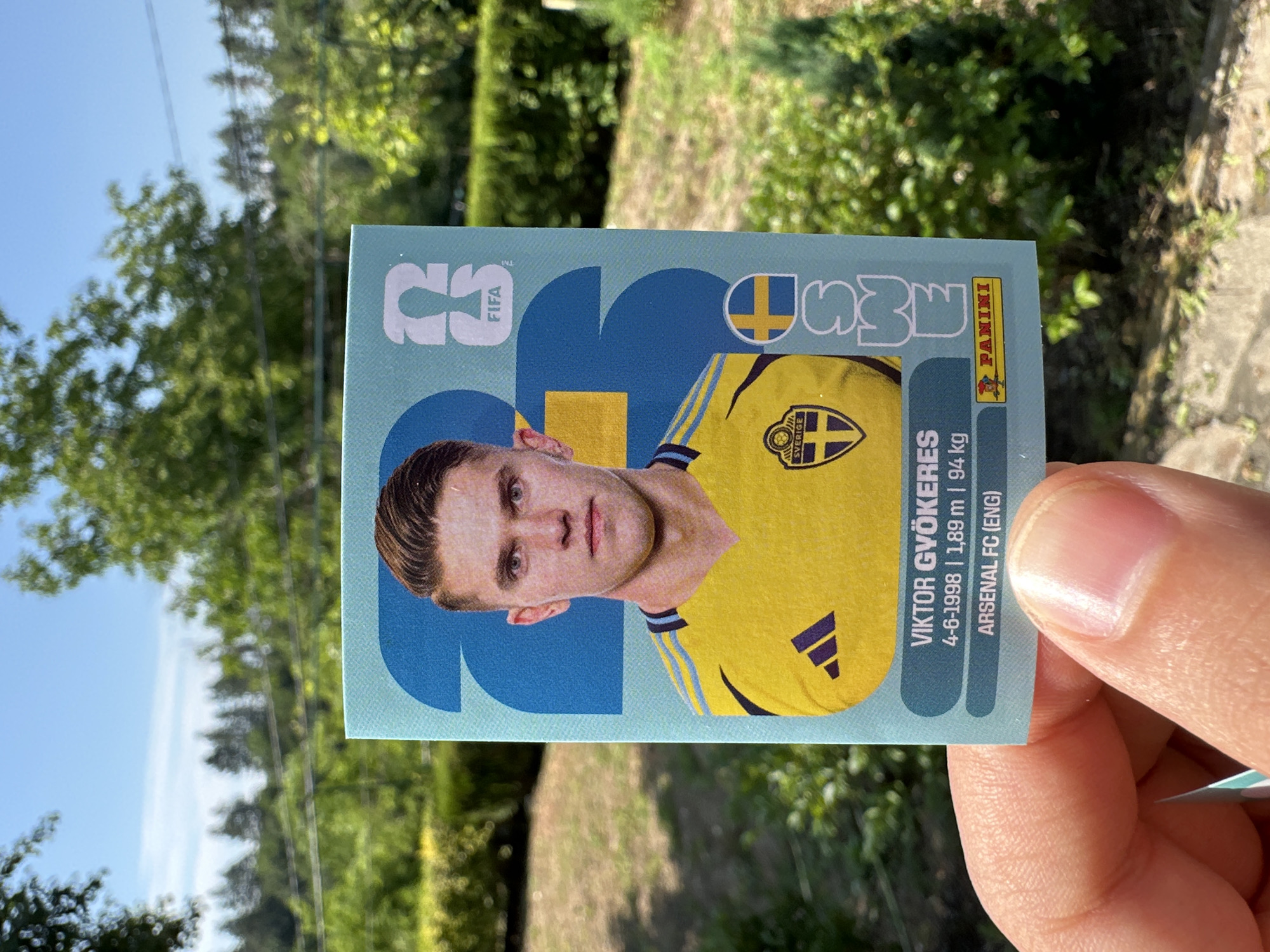 A person's hand holds up a trading card featuring a portrait of a male soccer player, Viktor Gyökeres, in a yellow Swedish national team jersey. The card has a light blue background with large white numbers and a FIFA logo. Text below the player's image includes his name, date of birth, height, weight, and club, "ARSENAL FC (ENG)". The card also displays the Panini logo. The background of the image shows a blurred scene of green trees and foliage under a bright blue sky, suggesting an outdoor setting, possibly in Prisovo, Bulgaria.