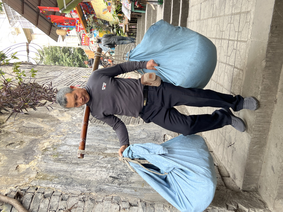 A man in a gray long-sleeved shirt and black pants balances two large blue sacks on a bamboo pole across his shoulders. He stands on a gray stone pathway next to a stone wall with greenery above. A tan pouch is attached to his belt. Shops and people are visible in the background. The image is rotated clockwise.