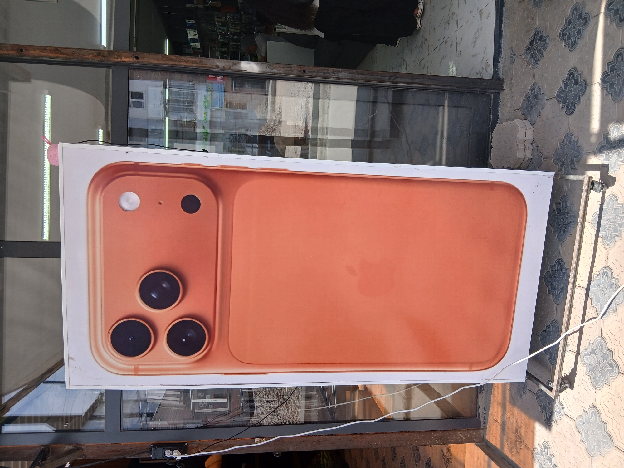 A large, vertical advertisement for an iPhone is displayed on a white frame positioned outside a shop. The advertisement features a close-up of the rear of a gold-colored iPhone, showcasing its triple-lens camera system and an Apple logo. The shop appears to be a mobile phone store, with shelves of phones visible through the glass doors. The advertisement is situated on a tiled sidewalk in front of the shop. The scene is outdoors, with natural lighting.