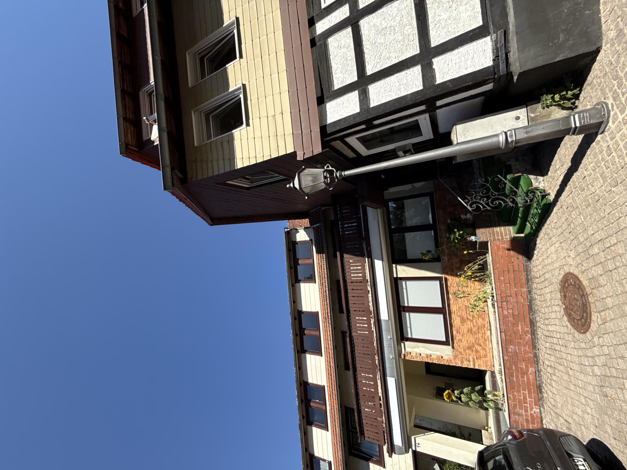 This is a low-angle, wide shot of a street in Bad Sachsa, Germany, with a clear blue sky. The foreground is dominated by a cobblestone street and a parked car. To the right, a distinctive half-timbered building with dark wood beams and white infill is visible, angled upwards. A traditional black street lamp stands prominently in front of this building, casting a shadow onto the cobblestones. Further left, another building with a cream-colored facade and a wooden balcony is seen. A single tall sunflower stands near the entrance of this building. The scene appears to be a residential street on a sunny day.