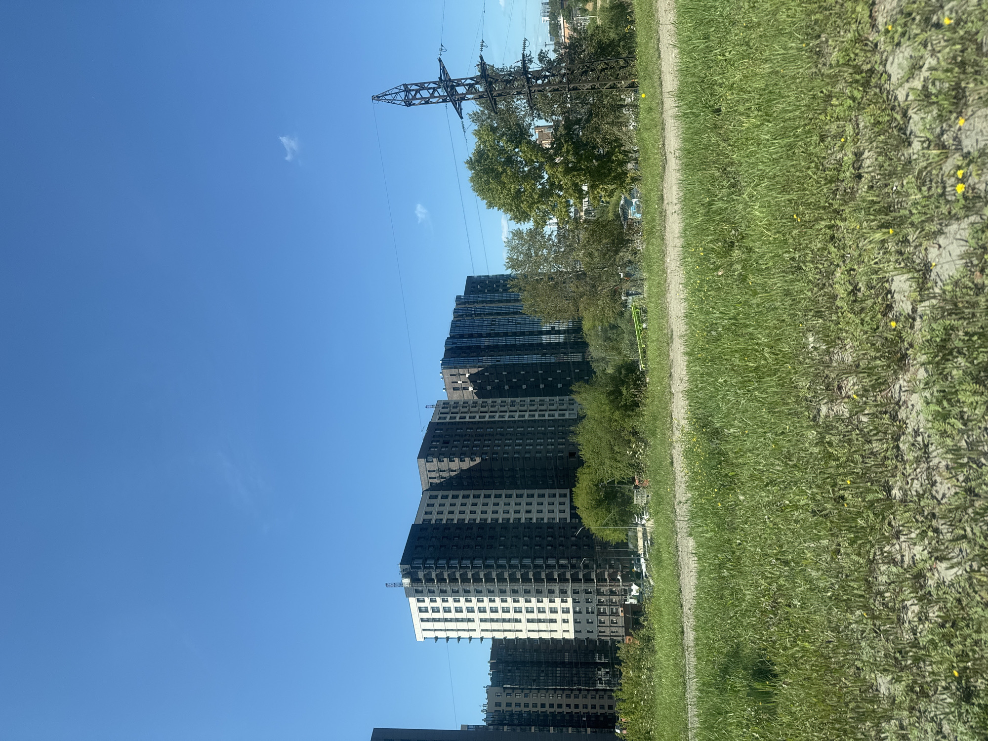 The image shows a row of modern, multi-story apartment buildings under a clear blue sky. The buildings have a mix of light and dark facades, with many windows visible. In the foreground, there is a field of green grass with some sparse yellow wildflowers. To the right of the buildings, a tall, dark metal power pylon with power lines is prominent. Behind the buildings, more trees and some distant structures can be seen. The scene appears to be an urban or suburban area on a sunny day. Based on the architectural style of the buildings and the presence of the power pylon, this scene is likely located in a newer development area of Irkutsk, Russia, possibly on the outskirts of the city. There are no people visible in the image.