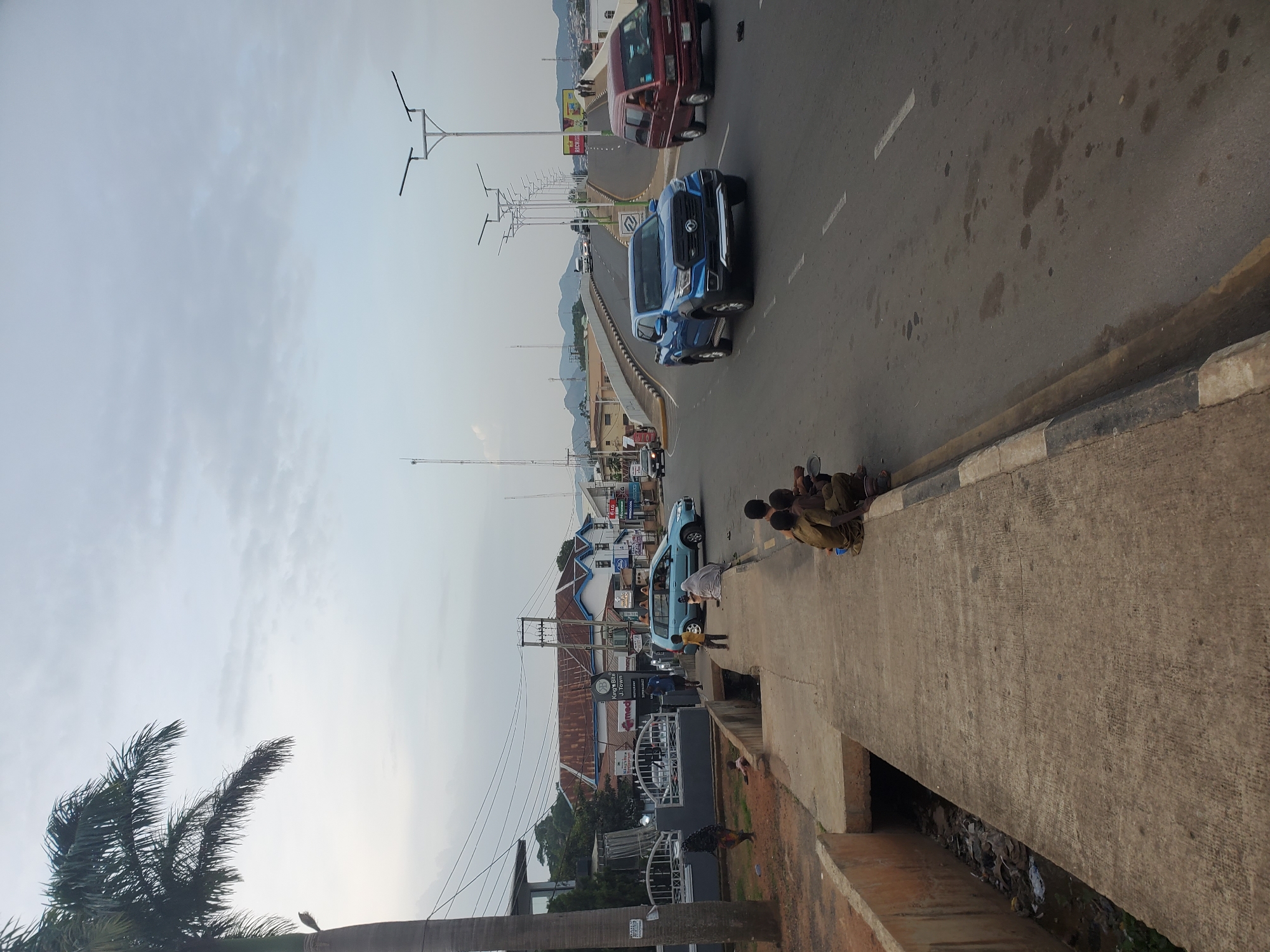 The image captures a street scene in Jos, Nigeria, focusing on a busy road and its surrounding environment. In the foreground, a concrete walkway runs alongside a drainage ditch filled with debris and litter. Several children are seated on the edge of the walkway, appearing to be interacting with each other. A palm tree is visible on the left side of the image, its fronds extending towards the overcast sky.
The road itself features a modern overpass with streetlights and traffic. A blue pickup truck is in motion on the right side of the road, while a red van is also visible. Several other vehicles, including a light blue sedan, are seen on the overpass and in the background. Buildings line the street, some with visible signage, indicating a commercial area. The overall atmosphere suggests a typical urban environment in Jos, with signs of development and everyday life.
