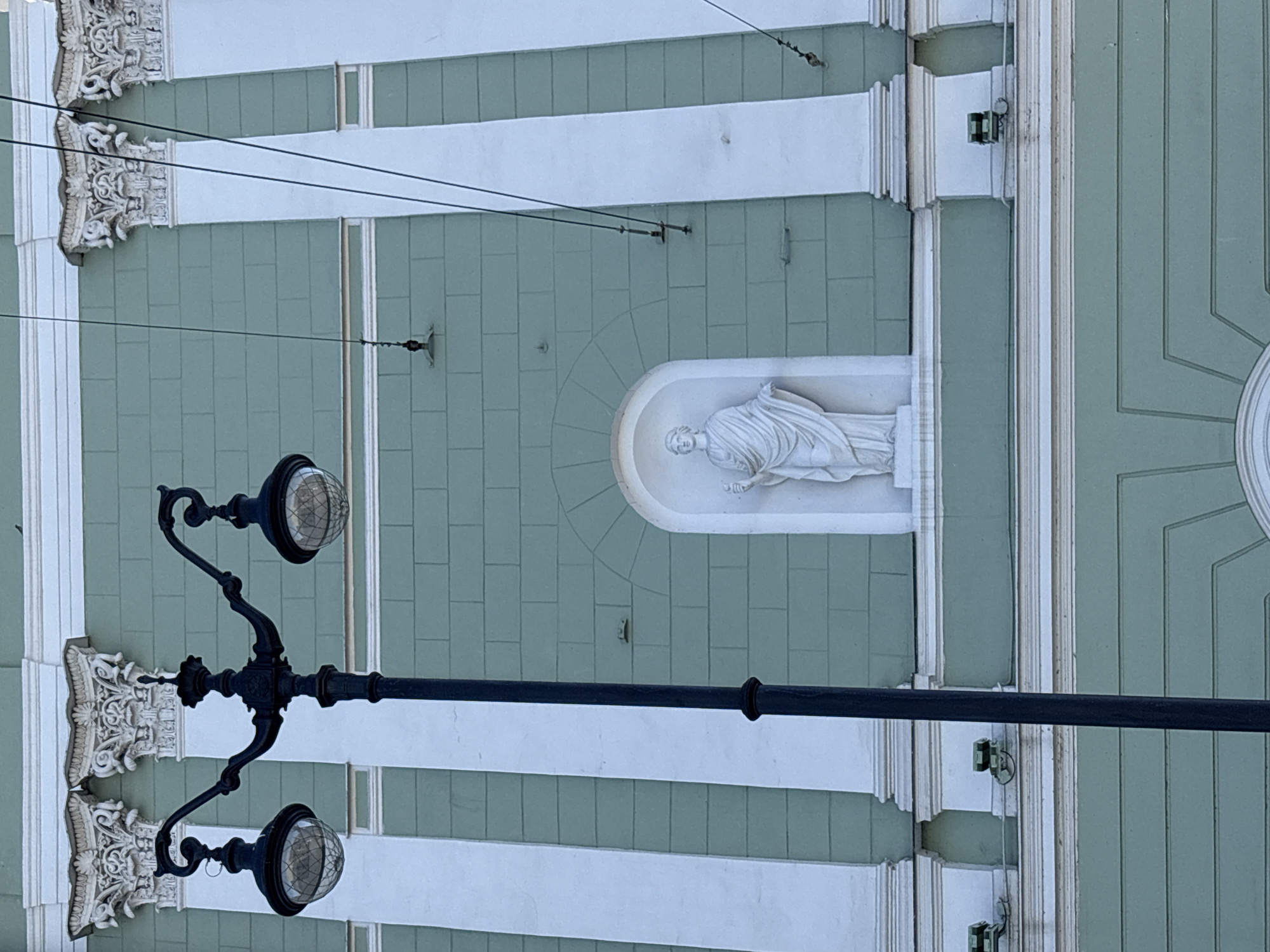 The image captures a detailed, upward-angled view of a building facade, characteristic of Saint Petersburg, Russia, partially obscured by a street lamp. The building features a light, muted green exterior with recessed horizontal lines suggesting a block pattern, juxtaposed with prominent white Neoclassical architectural elements. These elements include a classical cornice at the top, vertical pilasters or engaged columns adorned with ornate Corinthian capitals, and multiple horizontal bands and ledges.
Centrally within the frame, an arched niche is deeply recessed into the green wall, housing a full-length white marble statue. The statue depicts a draped classical figure, possibly male, holding an elongated object, possibly a scroll or staff, in its left hand, with its right arm slightly extended.
In the foreground, a black, ornate street lamp stands slightly left of center, featuring a central pole from which two curved arms extend horizontally, each supporting a spherical, glass-domed lantern with an internal grid pattern. Multiple thin black overhead wires, indicative of trolleybus or tram lines, traverse the scene, attached to the building facade at various points by dark metal brackets and insulators. Several small, dark green rectangular objects, potentially security cameras or external lighting fixtures, are mounted on the white horizontal ledges in the lower section of the facade. A single small bird is perched on the upper white cornice, above the rightmost Corinthian capital. The scene is brightly illuminated by natural daylight.