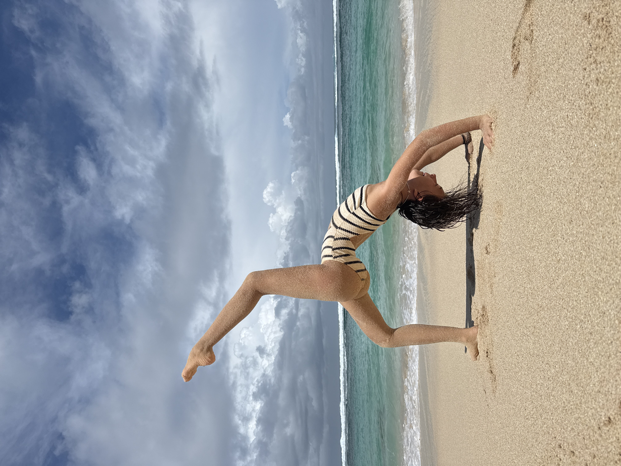 The image is a portrait-oriented photograph, rotated such that the sky is on the left and the beach and ocean are on the right, capturing a woman performing a flexible yoga or gymnastic pose on a sandy beach in Kuta Selatan, Indonesia. The subject, a woman, is positioned diagonally across the frame, in a deep backbend. Her body, partially covered in golden sand, is clad in a striped, possibly beige and dark-colored, one-piece swimsuit. Her dark, wet hair is splayed on the sand behind her head as she stretches, with hands seemingly supporting her on the sand and one leg extended upwards.
The scene depicts a beautiful beach environment. The golden-brown sand of the beach is prominent on the right side of the rotated image. Adjacent to the sand is the clear, light blue and turquoise water of the ocean, with subtle white waves breaking along the shoreline. The horizon line of the ocean is visible. The sky, filling the left side of the frame, is partly cloudy, displaying patches of bright blue interspersed with soft white and more substantial grey clouds. The lighting indicates it is daytime. No discernible text is present within the image.