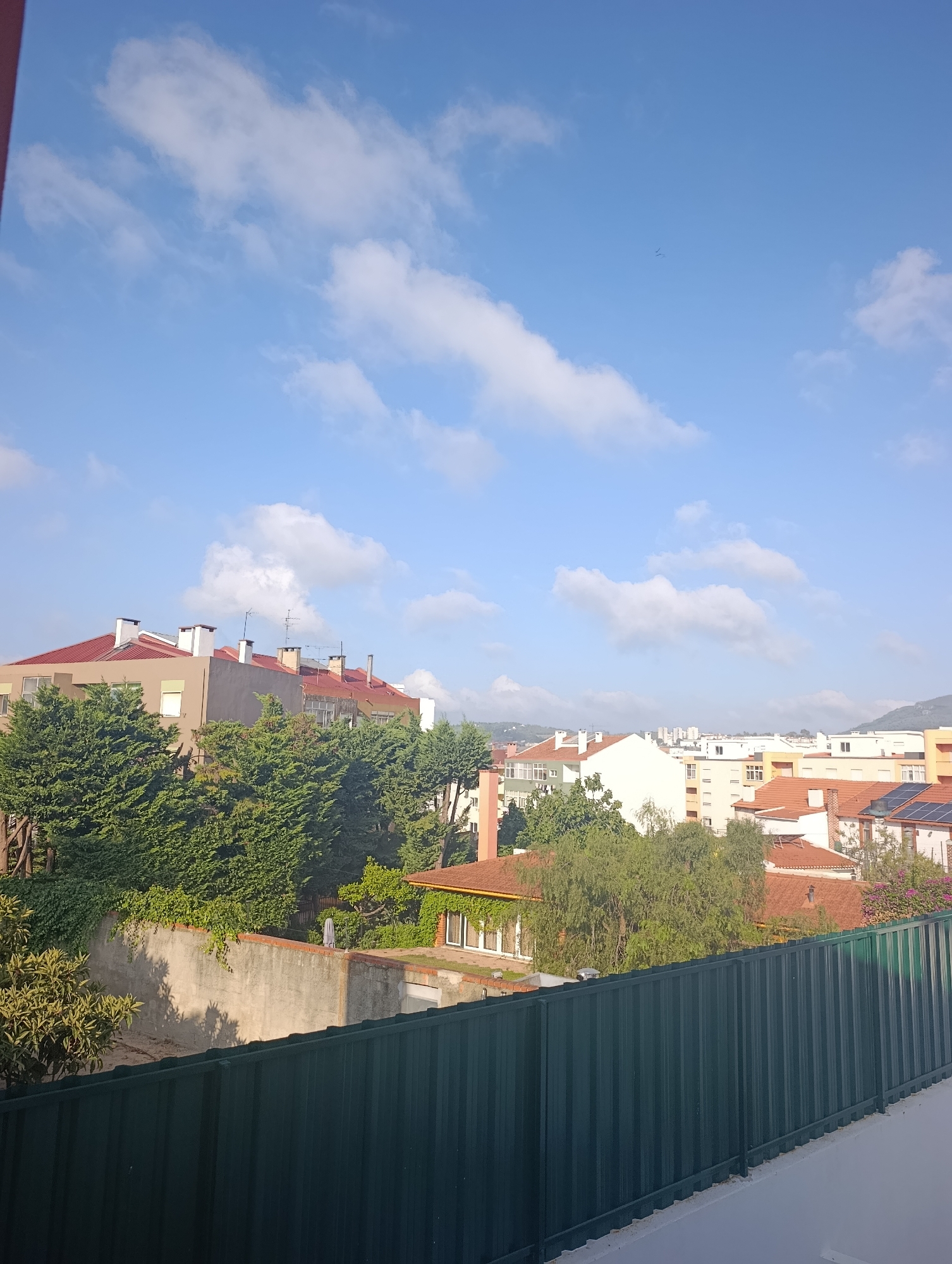 The image captures a sunlit, urban cityscape in Setúbal, Portugal, under a blue sky dotted with wispy white clouds. The foreground is dominated by a dark green corrugated metal fence, suggesting a balcony or rooftop vantage point. Beyond the fence, a series of residential buildings with terracotta tiled roofs and light-colored facades are visible, interspersed with lush green trees. Some buildings feature satellite dishes and solar panels, indicating modern amenities. In the distance, more buildings and a hazy, green hillside can be seen. Two small dark birds are visible high in the sky, appearing as tiny specks. The scene appears tranquil, with no people or immediate activity evident. The overall impression is a typical residential area bathed in daylight.