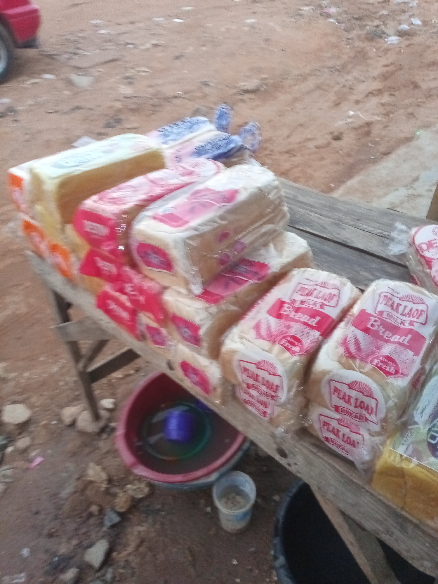 A slightly blurry, eye-level, full shot shows a collection of packaged loaves of bread displayed on a rustic, weathered wooden table. The bread loaves are wrapped in clear plastic and feature prominent red and white labels that read "PEAK LOAF MILK BREAD Always Fresh". Some of the loaves are stacked in multiple layers. The table is set on a dusty, reddish-brown dirt ground, characteristic of an outdoor market or roadside stall. In the background, out of focus, is a red vehicle and a sandy, uneven terrain with scattered debris. A red plastic basin containing water and a blue object is visible on the ground to the left of the table, and a small clear plastic cup with some contents sits near the basin. The overall scene suggests a humble, local commerce setting, likely in a part of Jos, Nigeria where such informal trading is common, with the environment appearing somewhat unkempt and typical of such areas.