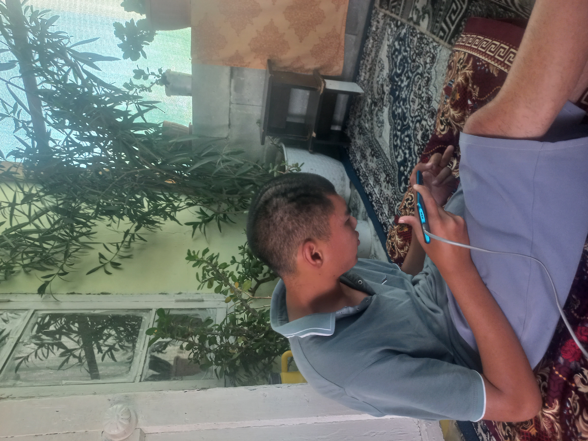 The image depicts a young person sitting on a patterned rug, engrossed in a mobile phone. The person is wearing a gray polo shirt and light blue shorts. A charging cable is connected to the phone. The environment appears to be an outdoor or semi-outdoor space, possibly a veranda or patio, with a window with white framing visible in the background, reflecting green foliage. Several potted plants are scattered around, some in white pots and others in terracotta. A small wooden stool is placed to the right, next to a wall covered with a patterned fabric. The ground is covered with a large, ornate rug. The overall scene suggests a relaxed, domestic setting. The location is identified as Бакмал, Kyrgyzstan.