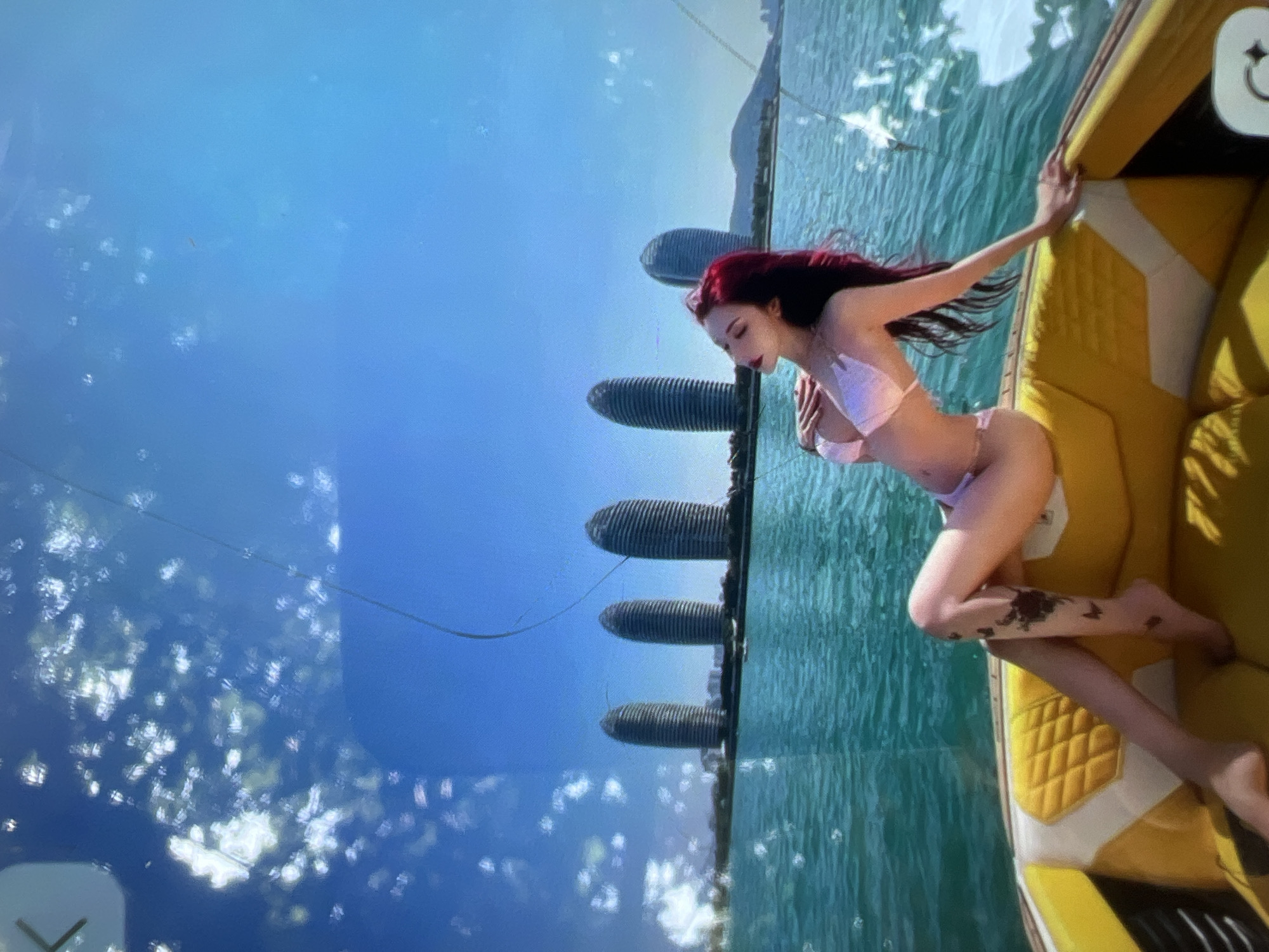 A woman with long, dark red hair is seated on the edge of a yellow and white upholstered boat, wearing a pink bikini. She is leaning back with one hand resting on her chest and the other extended towards the side of the boat. Her legs are crossed, and she has dark tattoos on her left leg. The boat is on a bright turquoise body of water, with a clear blue sky overhead. In the background, a distinctive line of tall, curved, ribbed buildings rises from the water's edge, with a hazy mountainous landscape beyond. The image appears to have some light reflections or disturbances on its surface, particularly in the upper and right portions. The setting does not resemble the city of Kara-Dıykan, Kyrgyzstan, which is an inland city and not located near such a coastal or large body of water with this type of architecture.