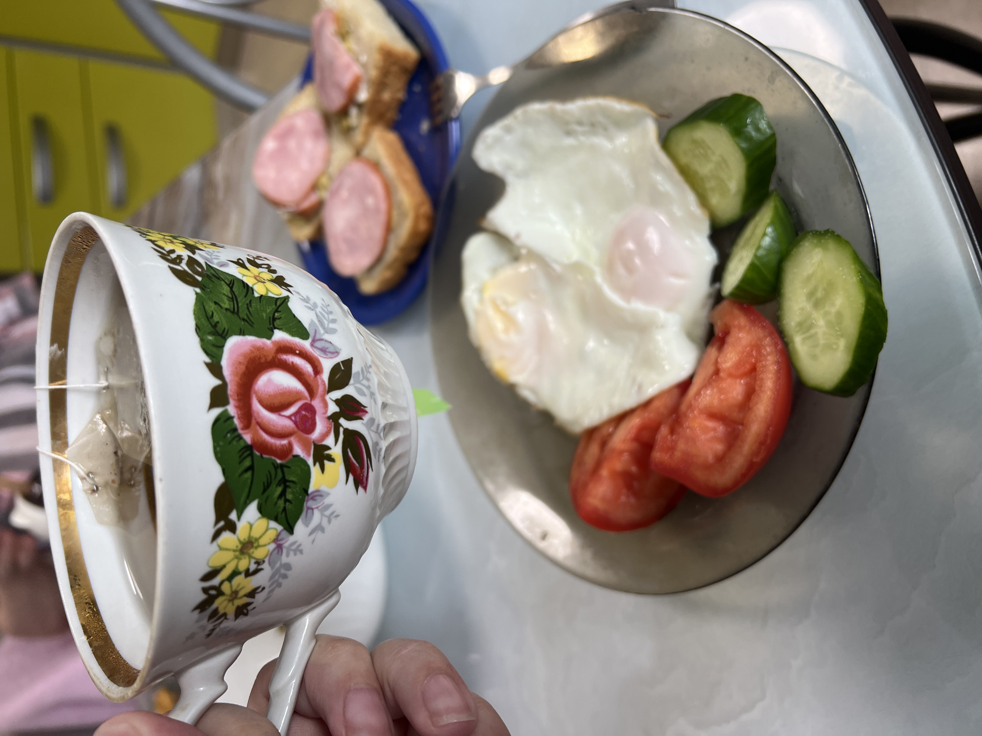 A photograph shows a breakfast scene in Perm, Russia. A person holds a floral-patterned teacup with a gold rim, containing a teabag. Next to the cup is a gray plate with two fried eggs, sliced cucumbers, and sliced tomatoes. A blue plate with bread and sausage slices is partially visible in the background. There is a fork on the blue plate. A yellow cabinet and metal bars are also visible in the background. The photograph seems to be taken indoors on a marble-like surface.