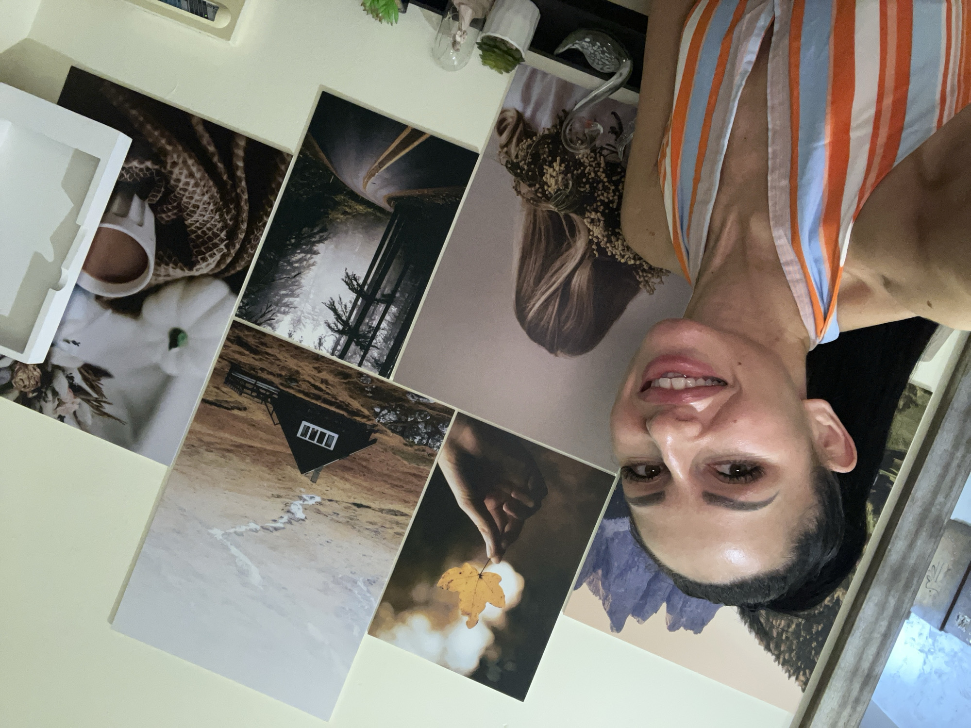 The image displays a collection of printed photographs arranged on a light-colored wall. The photographs depict various scenes and objects. One image shows a close-up of a white mug with a brown liquid, placed on a textured brown blanket, with a white pumpkin and a sprig of white flowers next to it. Another photograph features a dark, misty forest with trees and a winding road. A third image shows a person's blonde hair adorned with dried flowers. There is also a picture of a small, dark cabin with a white-framed window, situated on a hill with a light-colored path leading to it, amidst a rocky, sparse landscape. Another photograph shows a hand holding a single yellow leaf. In the background, a mountainous landscape with rugged peaks is visible.
The most prominent subject is a woman whose face is partially visible at the bottom right of the image, positioned upside down relative to the other photographs. She has dark hair pulled back, brown eyes, and is smiling with her teeth showing. She is wearing a striped top with vertical bands of orange, white, and blue.
The overall impression is one of personal decor, with curated images chosen for display. The photographs suggest a theme of nature, coziness, and perhaps travel. The lighting suggests an indoor setting, likely daytime, given the brightness of the wall. There is no visible text in the image.