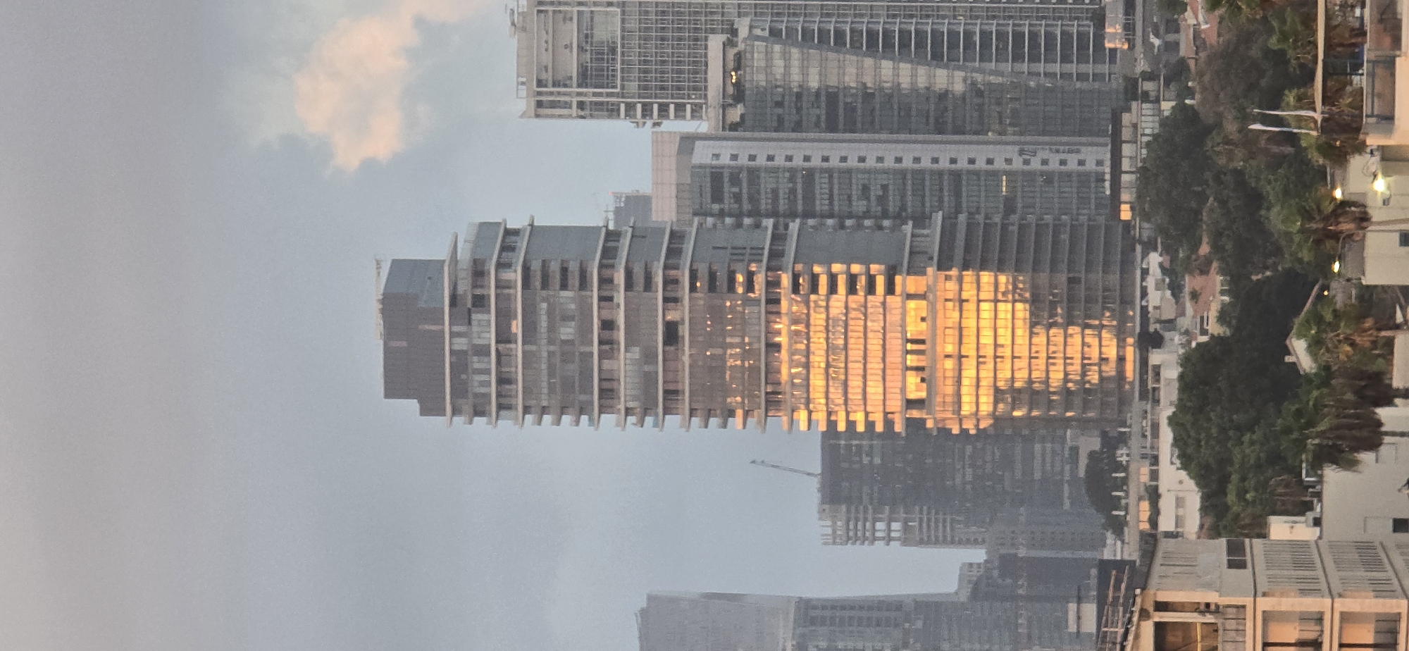 The image depicts a cityscape with a prominent modern skyscraper reflecting the warm golden light of the setting sun. The skyscraper is the central focus, characterized by its tiered structure and glass facade. Adjacent to it, other high-rise buildings of varying architectural styles are visible, some appearing to be under construction. In the foreground, lower-rise buildings and dense green trees, including palm trees, create a contrast with the towering structures. The sky is overcast with a hint of soft pink clouds, suggesting either early morning or late evening. The scene is consistent with the urban landscape of Tel Aviv, Israel, known for its contemporary architecture and coastal setting. No people are visible in the frame.