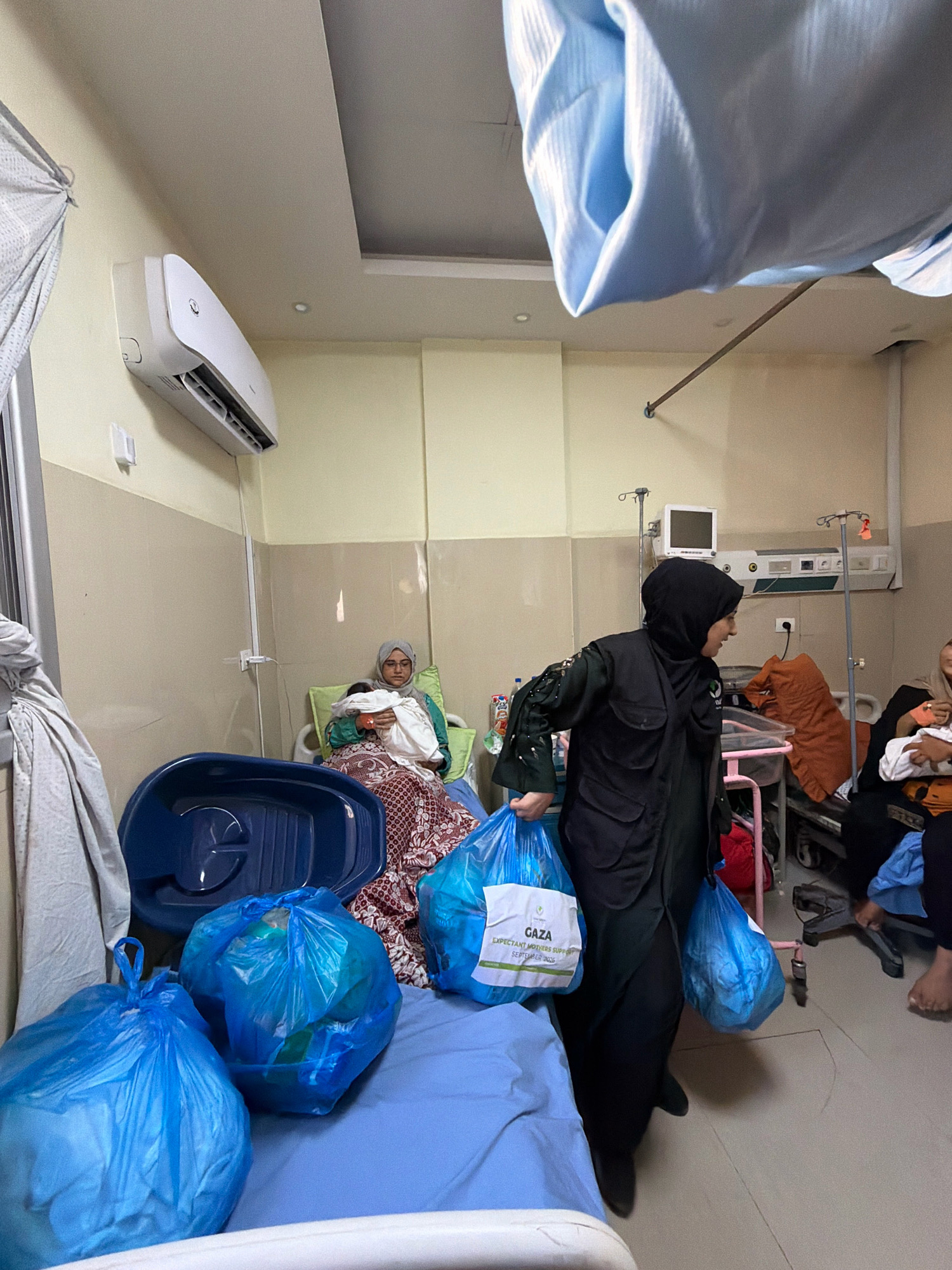 In a hospital room in Gaza, Palestinian Territories, a woman wearing a black hijab and vest is seen from the waist down carrying two large blue plastic bags filled with supplies. A white label on one bag reads "GAZA EXPECTANT MOTHERS SUPPORT SEPTEMBER 2023". In the background, a woman with a gray hijab sits on a hospital bed, holding a newborn baby wrapped in white cloth. The room contains medical equipment including an air conditioning unit, an IV stand, and a monitor. Several blue plastic bags are also on the hospital bed.