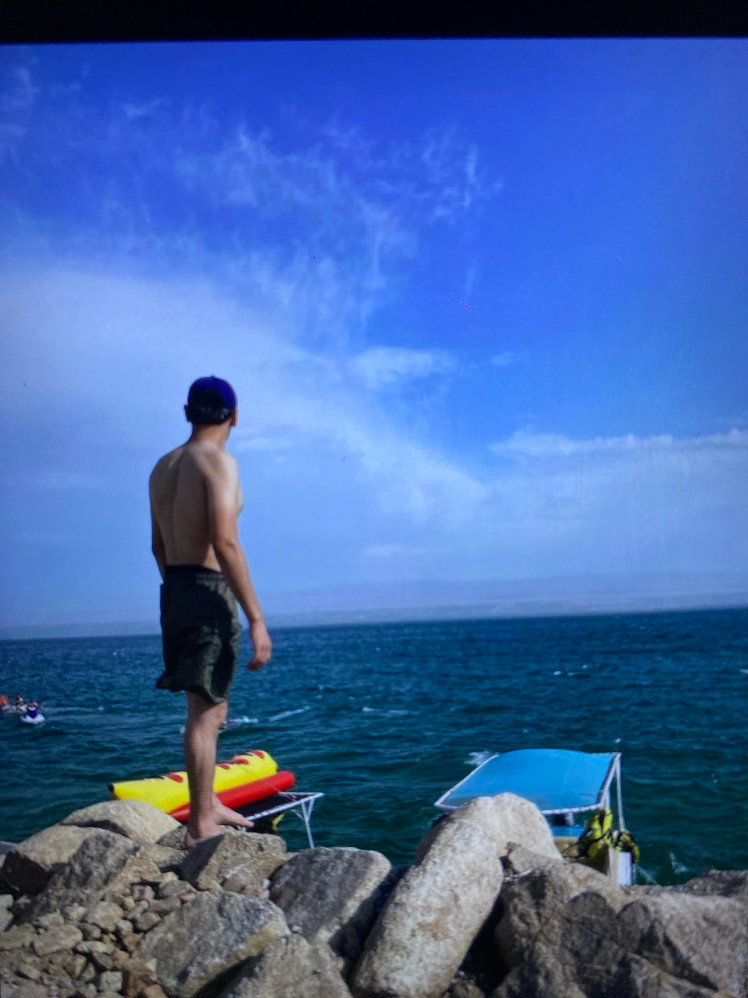 The image depicts a man standing on a rocky shore overlooking a body of water. He is shirtless, wearing a blue baseball cap and dark shorts. In the water, there is a yellow and red inflatable tube and a small boat with a blue canopy. The sky is blue with scattered clouds. The scene is set during the daytime. The location is not identifiable as Ramenskoye, Russia, as no specific landmarks or contextual information is present to suggest this location.