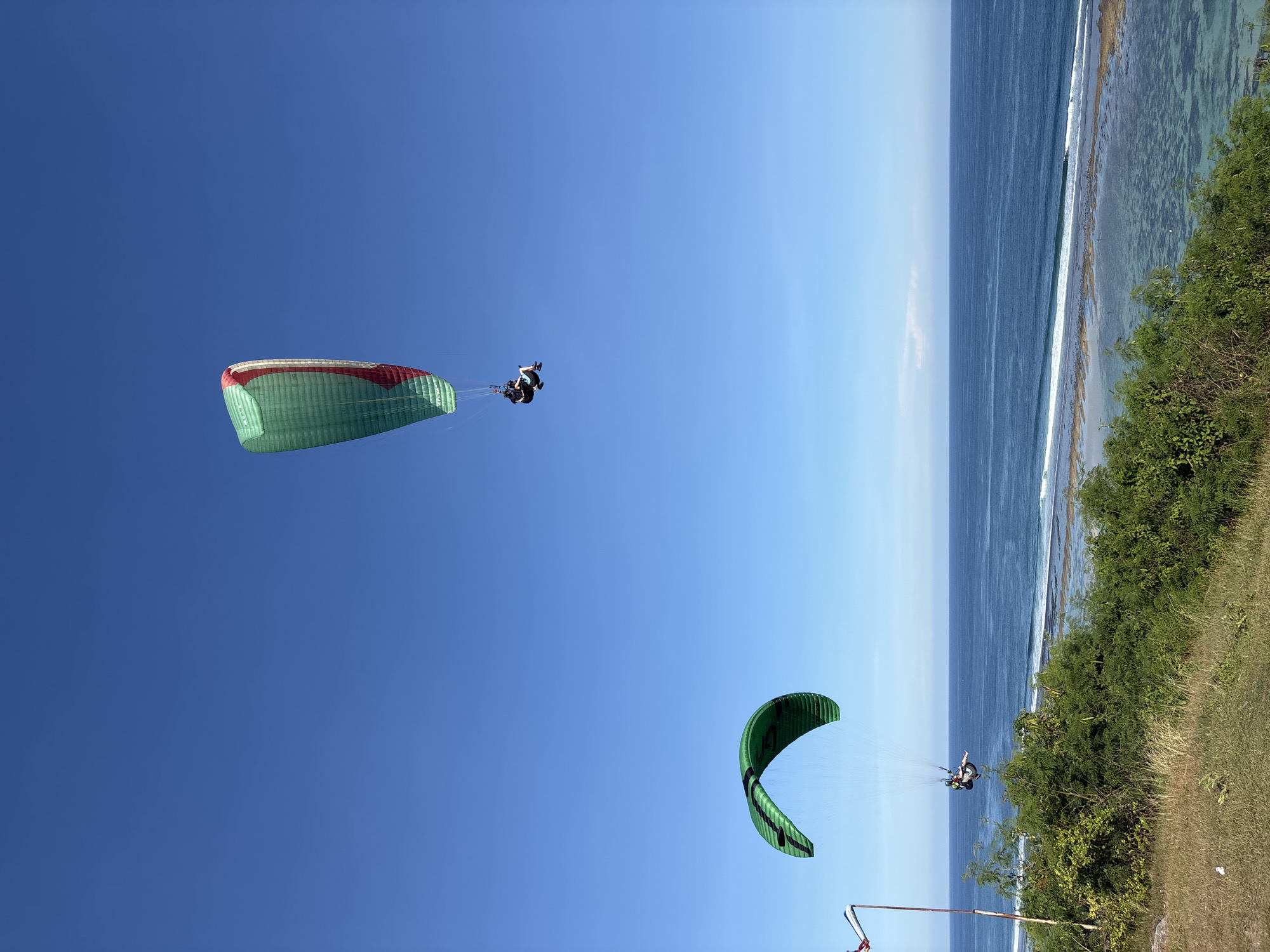 Two paragliders are airborne above the coast of Kuta Selatan, Indonesia. The upper paraglider is primarily green with red accents, and a person is suspended beneath it. The lower paraglider is green, and a person is suspended beneath it as well. The sky is clear and blue. The ocean borders the coastline, and some foliage is visible along the shore. A pole and some other objects are at the bottom.