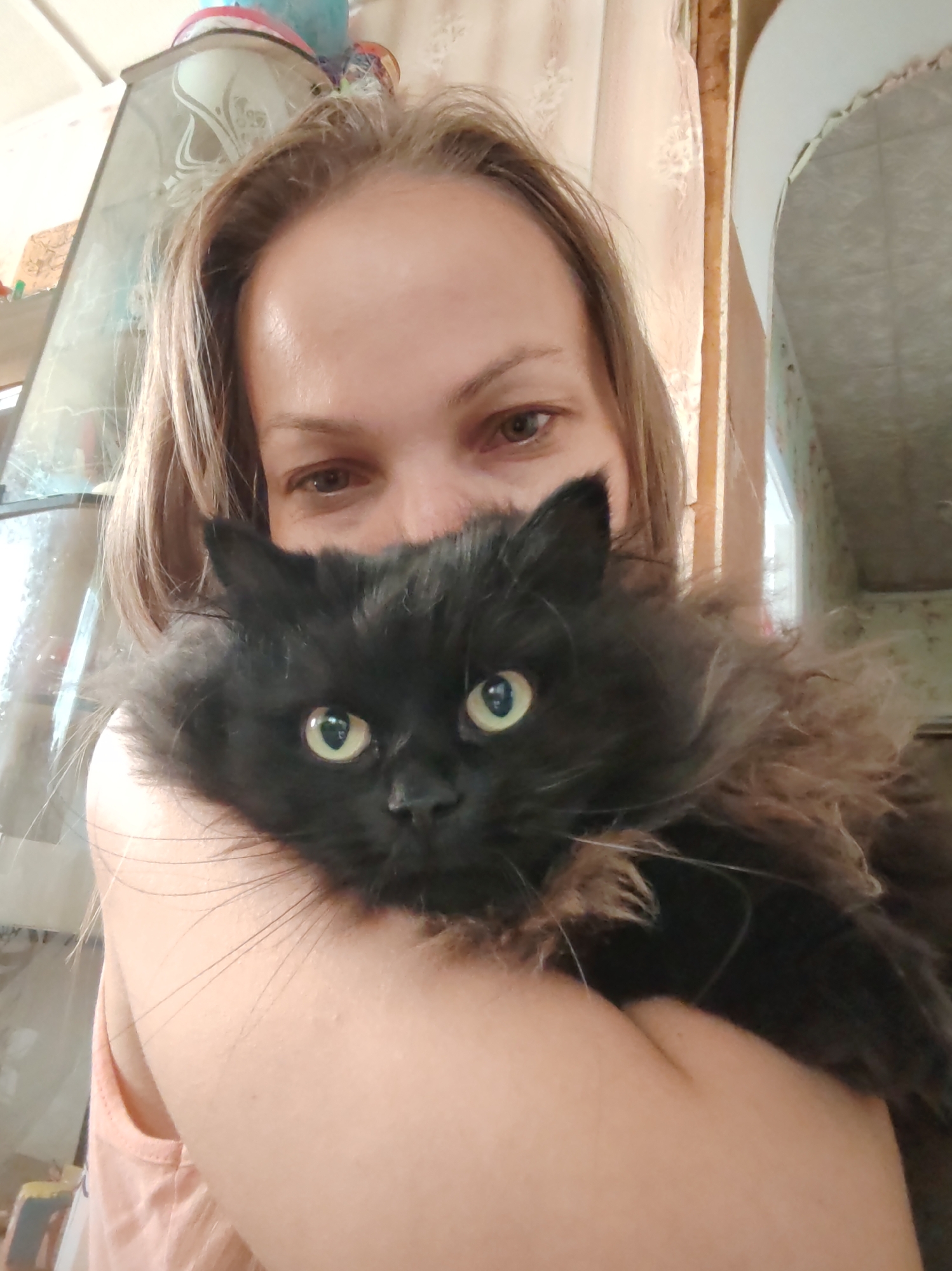 A close-up, selfie-style portrait shows a woman holding a fluffy black cat. The woman's face is partially obscured by the cat and her hair, but her eyes are visible and appear to be light brown. She is wearing a peach-colored tank top. The cat has bright green eyes and is nestled against the woman's chest. The background is blurred, but suggests an indoor setting with furniture and curtains. The scene is likely located in Chukhlem, Russia, though no specific landmarks are identifiable.