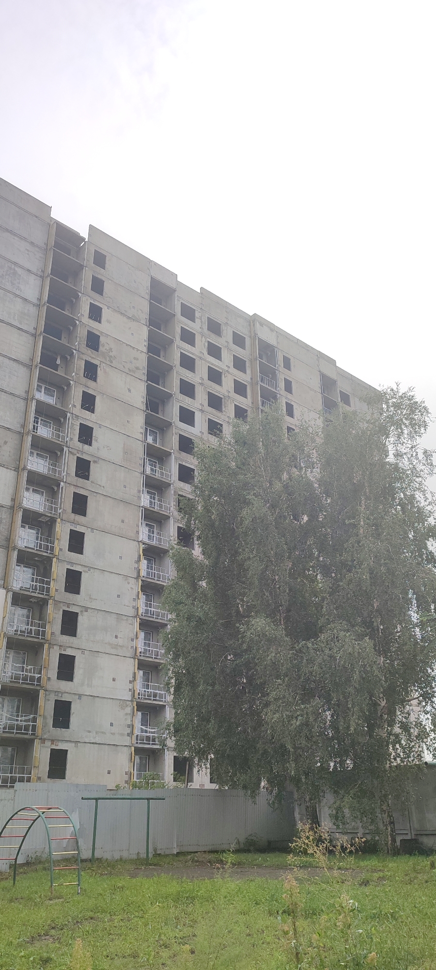 An unfinished concrete building stands tall against an overcast sky in Novoaltaysk, Russia. The building, appearing to be a multi-story apartment complex, has rows of empty window openings and balconies. A large, leafy tree partially obscures the facade of the building, its branches reaching towards the structure. In the foreground, a green playground climbing frame with red and yellow rungs is visible on a grassy area, along with a simple green metal horizontal bar. A light grey corrugated metal fence runs horizontally across the image, separating the playground area from the building. The overall scene suggests an abandoned or stalled construction site.