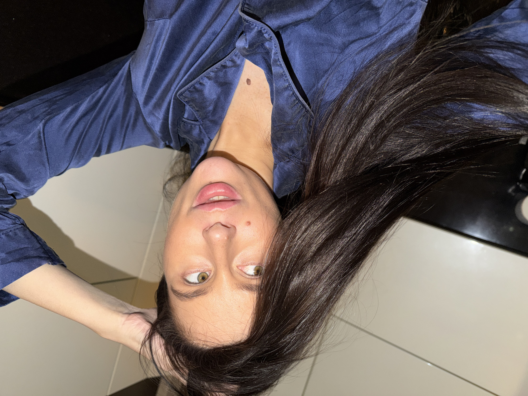 An upside-down portrait captures a young woman with dark, flowing hair. Her head is positioned at the bottom of the frame, looking upwards and slightly to her left. She has light brown or hazel eyes, and her lips are slightly parted, revealing a pink hue, possibly from lip gloss or lipstick. A small, dark brown mole is visible on her upper chest, between the lapels of her clothing. She is wearing a dark blue, silky shirt or pajama top, which has visible creases and a soft sheen. One arm is partially visible on the left, with her hand near her head, appearing to be resting or holding her hair.
The background is simple and appears to be an indoor setting, possibly in Khimki, Russia, as per the provided context. It consists of light-colored, possibly tiled or laminated surfaces towards the bottom and right, and darker, undefined areas towards the top left. The lighting is soft and even, suggesting artificial light or an overcast day indoors, without harsh shadows. Her expression is neutral and calm, perhaps pensive or simply gazing. There is no visible text in the image.