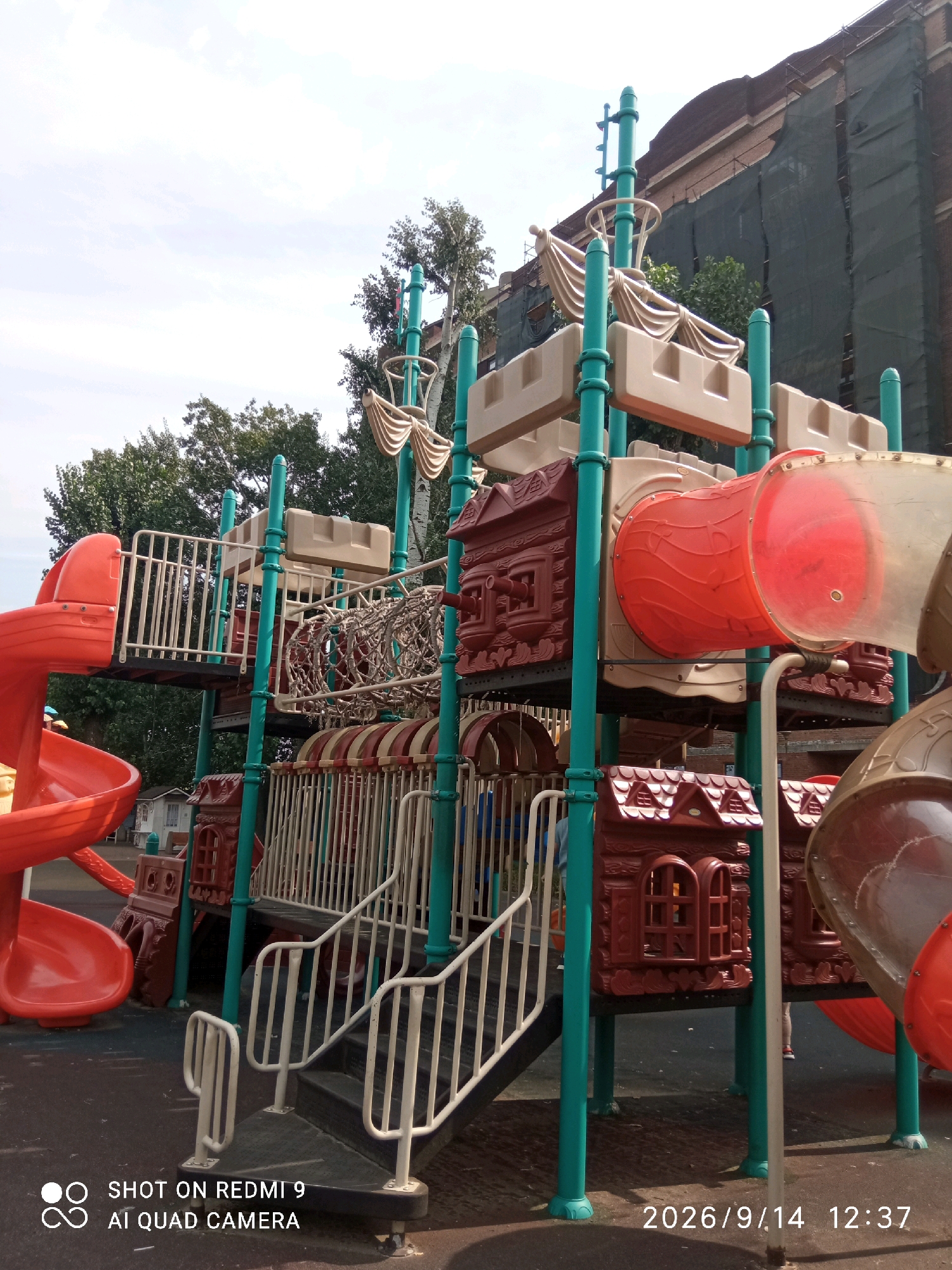 The image shows a playground on a cloudy day. The playground features a large, colorful structure designed to resemble a pirate ship. The structure includes red slides, tan turrets, and brown house-shaped elements with windows. Green poles support the various levels of the playground. In the background, a large building is visible, covered in green scaffolding and netting, suggesting ongoing construction or renovation. Trees with green foliage are also present. The ground is a dark, textured surface. In the lower left corner, text reads "SHOT ON REDMI 9 AI QUAD CAMERA". In the lower right corner, text reads "2026/9/14 12:37". No people are clearly visible or engaged in any specific actions within the frame. The scene is a typical urban playground environment.