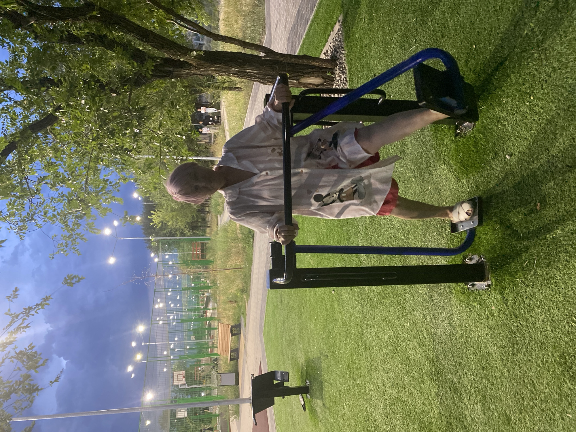 A middle-aged woman with short, dyed purple hair, wearing a long, white button-up shirt with Mickey Mouse graphics and red shorts, stands on an outdoor fitness machine in a park. The machine has black metal uprights and blue curved handles. The woman is positioned with her feet on the footplates of the machine and is holding onto the handles. She is looking towards the camera.
The park features artificial green turf flooring and a paved walkway in the background. Beyond the walkway is a chain-link fence enclosing a basketball court with lights. Trees with green foliage are visible, and the sky is dark blue with clouds and illuminated by artificial lights. The scene appears to be an outdoor recreational area, possibly a park or sports complex, in the city of Qarağandı, Kazakhstan. In the background, several other people are walking, and one person is pushing a stroller.