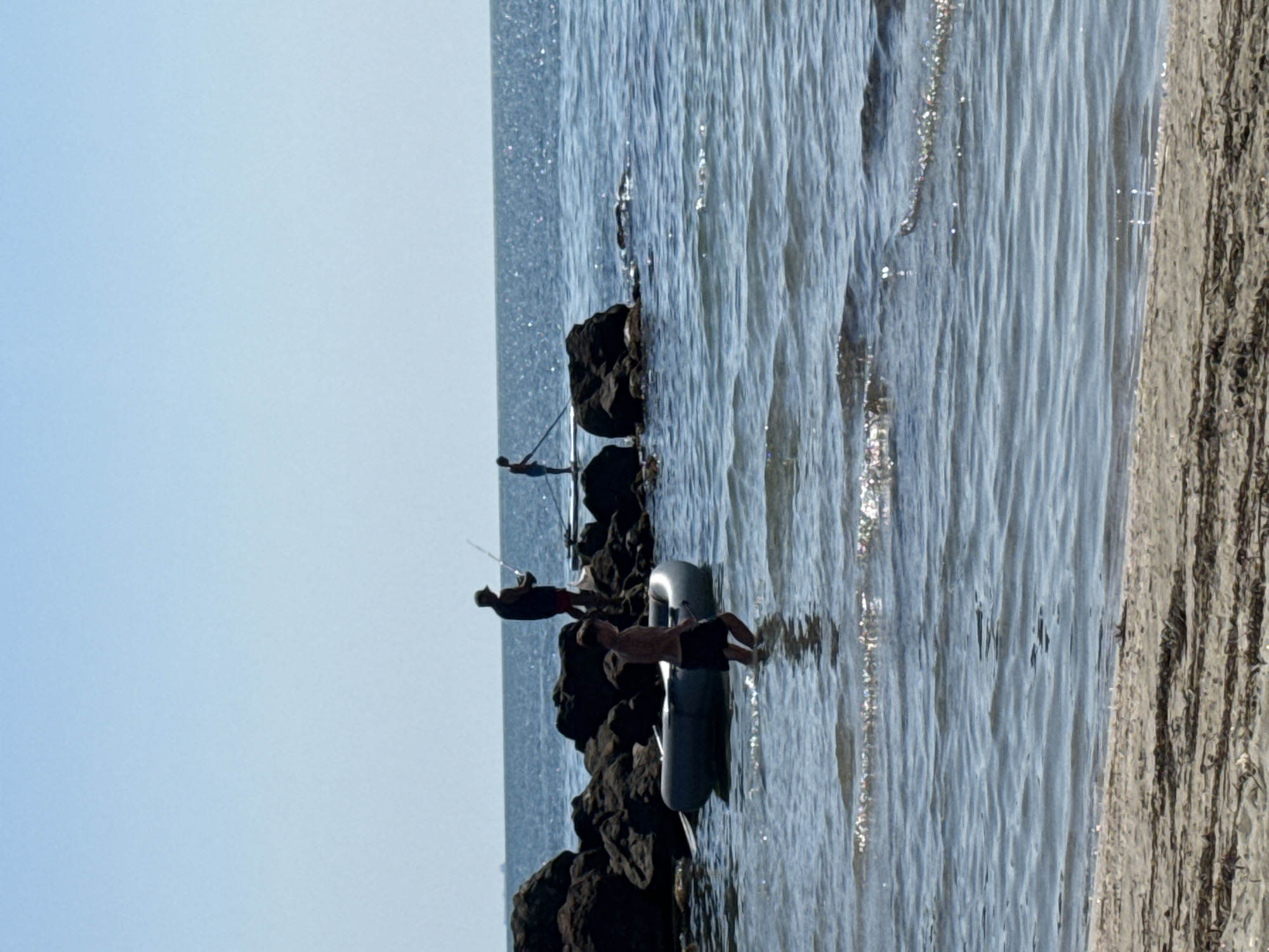 The image depicts a group of people engaged in recreational activities on a sunny day at the Pomorie beach, Bulgaria. In the foreground, a person is walking through shallow water, appearing to be heading towards a partially submerged inflatable boat. Further out, two individuals are positioned on rocks in the sea. One person, standing on the rocks, is holding a fishing rod, suggesting they are fishing. Another person is standing on a paddleboard, also in the water near the rocks. The background shows a clear blue sky and the vast expanse of the Black Sea. The beach in the very foreground consists of sand with some dried seaweed.