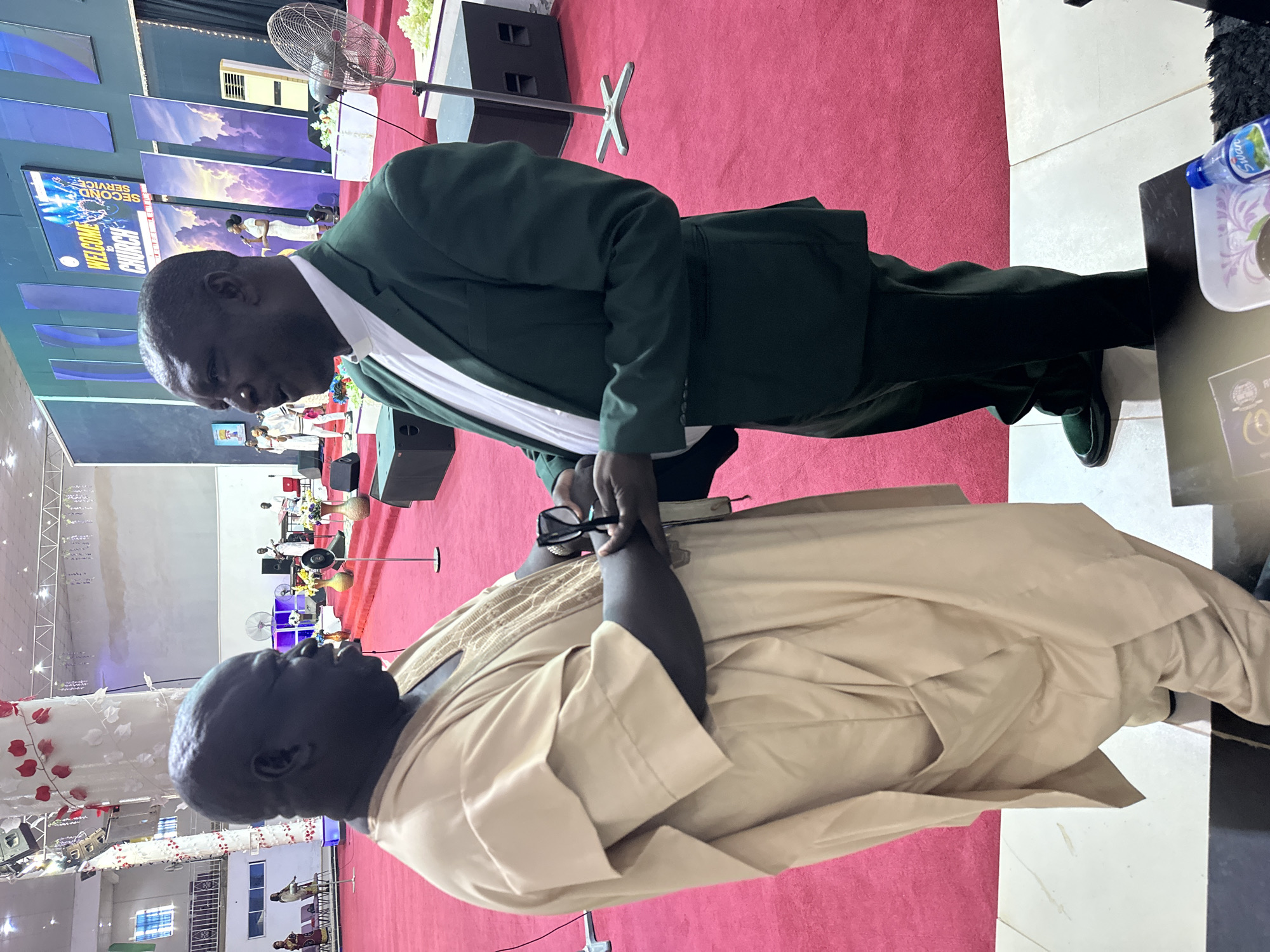 Two adult men are standing and interacting in a large indoor space with a red carpeted floor. The man on the left is wearing a light brown, long-sleeved robe with intricate gold embroidery around the neckline and chest. He has dark skin and is looking towards the other man. The man on the right is wearing a dark green suit jacket over a white shirt with a white collar, and dark green trousers. He also has dark skin and appears to be handing a pair of glasses to the man on the left. The background suggests a church or large event venue, with a stage visible in the distance featuring a screen with the text "WELCOME TO CHURCH" and "SECOND SERVICE". There are also sound equipment, fans, and floral decorations visible. The lighting appears to be artificial and bright, illuminating the entire scene. The environment is clean and well-maintained.