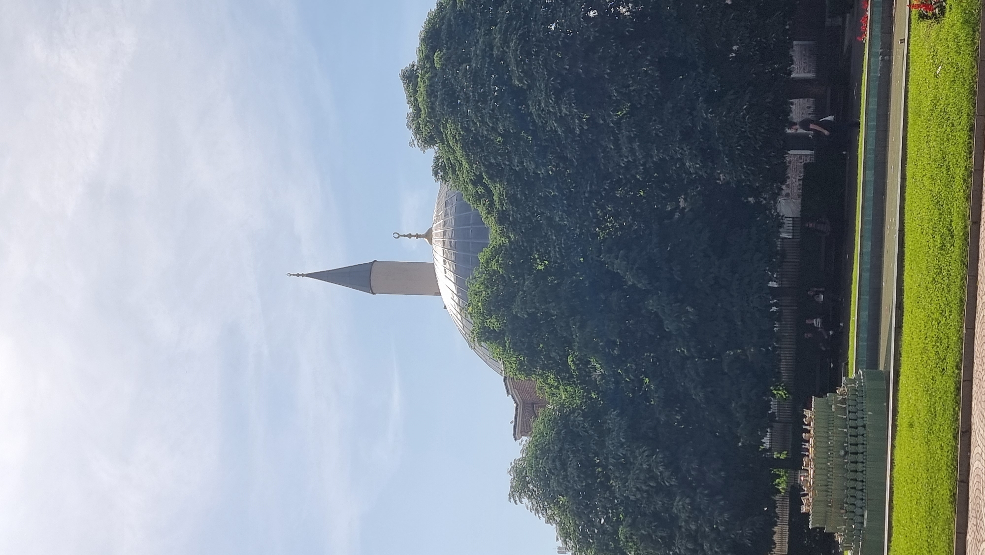 The image shows the Saint George Rotunda in Sofia, Bulgaria. The Rotunda's dome and a minaret can be seen partially obscured by trees. To the right, there is a park area with grass and a walkway, where some people are walking or sitting. The sky is blue with light clouds. The image appears to be taken from a low angle.
